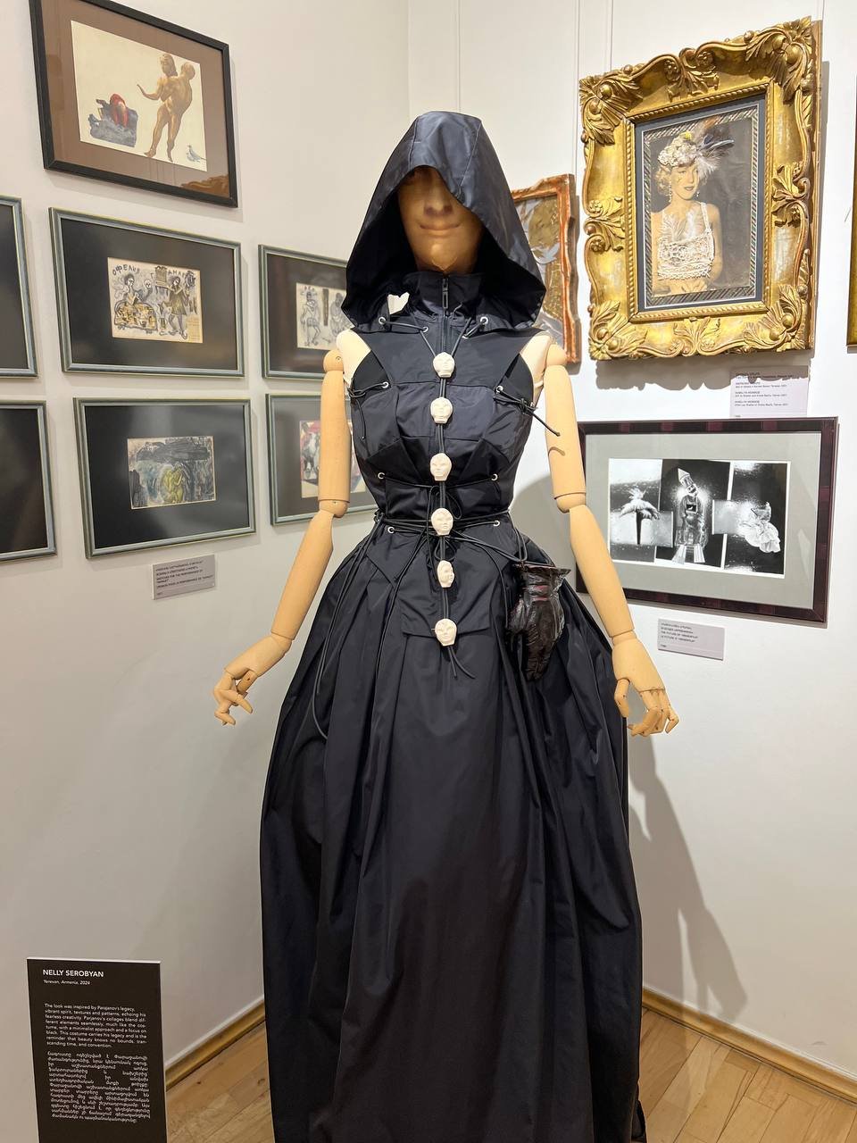 A mannequin dressed in a black, sleeveless, floor-length dress with a hood, displayed in an art gallery. The mannequin has wooden arms and a no face mask. It is surrounded by framed art photographs and drawings on the gallery wall.