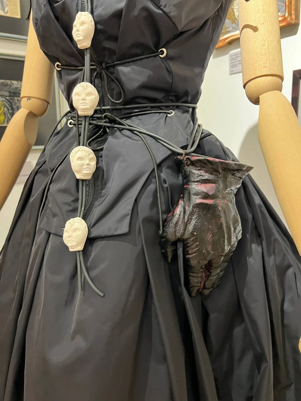 A mannequin dressed in black rain gear with a string of small, white, human face sculptures hanging from the waist and a large, dark, breast-shaped object attached to the skirt.