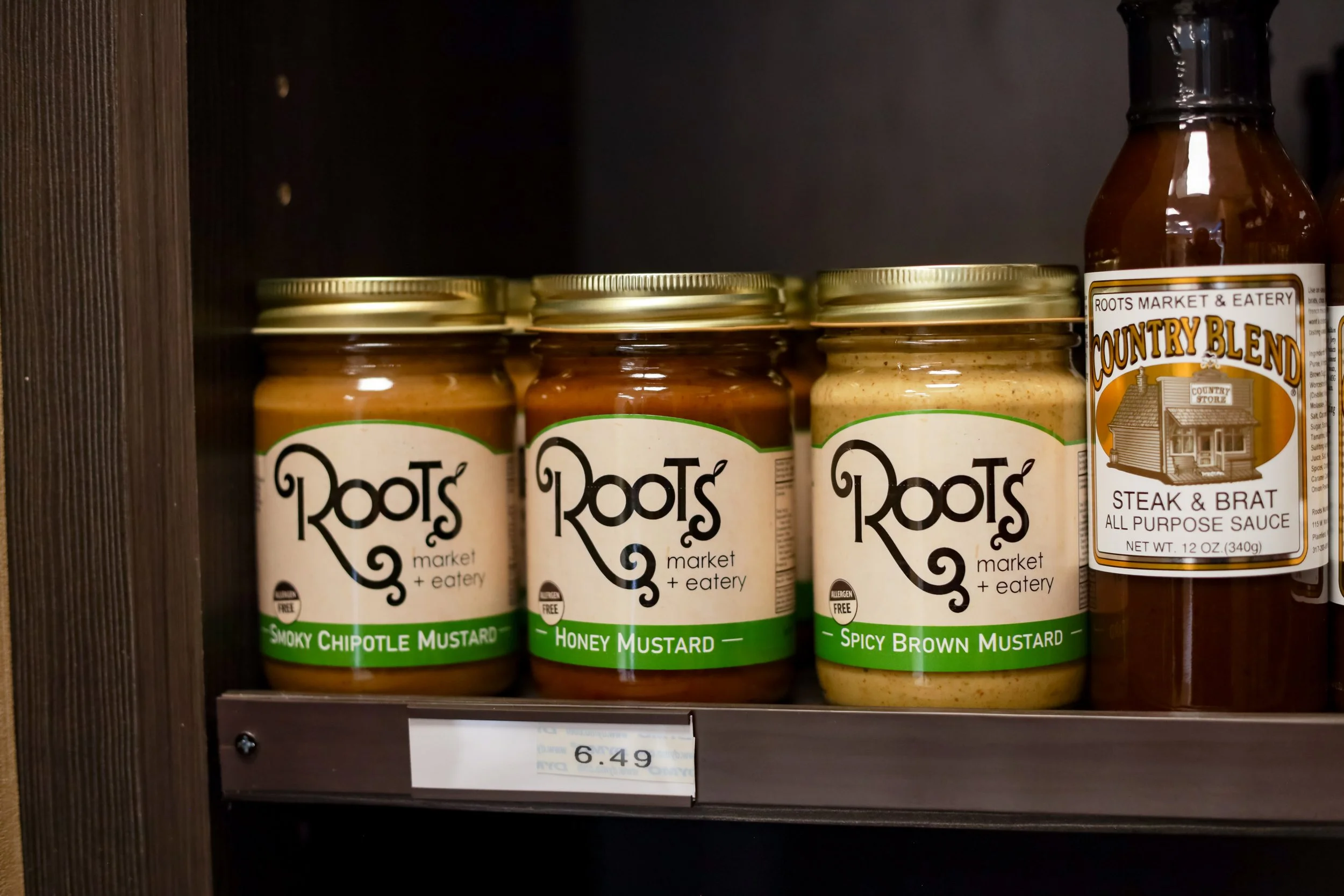 Three jars of Roots Market & Eatery mustards—Smoky Chipotle Mustard, Honey Mustard, Spicy Brown Mustard—on a store shelf, with a bottle of Country Blend Steak & Brat all-purpose sauce nearby.