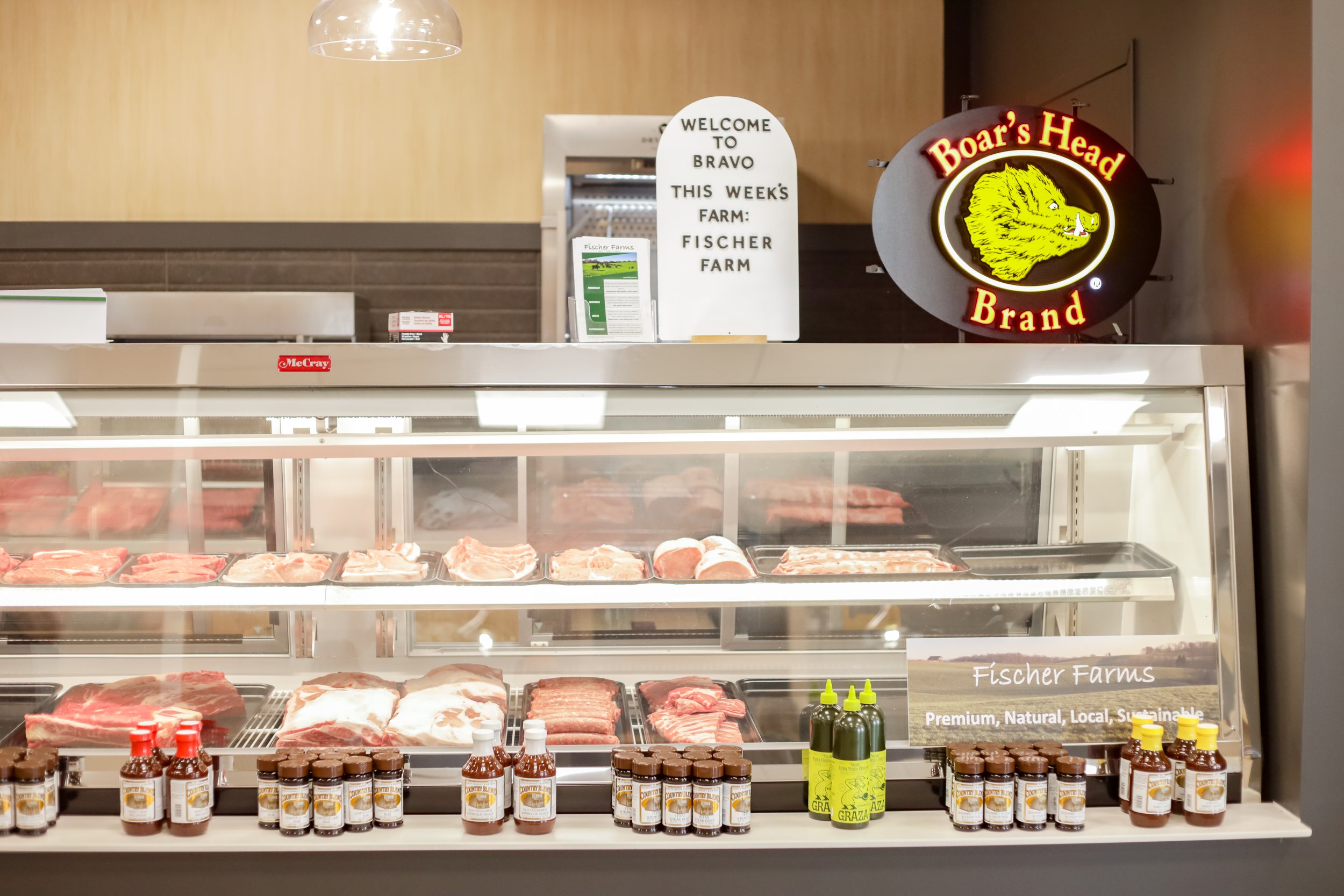 Meat display case at Bravo Butcher Shop with various cuts of meat and bottles of sauce, signs, and neon sign with a boar's head logo.