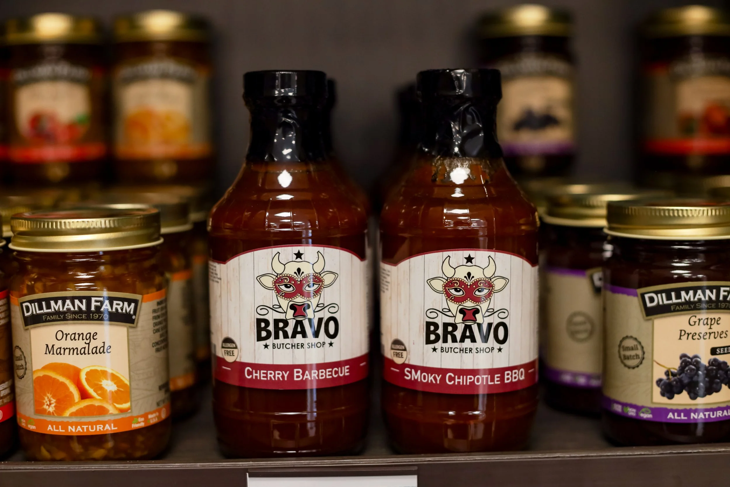 Shelf at Roots Market with sauce bottles, jars of orange marmalade, and grape preserves at a grocery store.