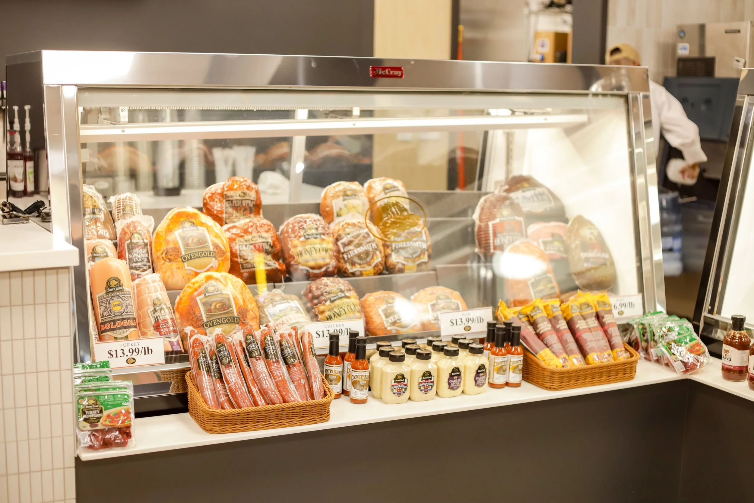 Display case at Bravo Butcher Shop with various meats including ham, sausages, and roast, priced at $13.99 per pound, with condiments and sauces on the counter in front.
