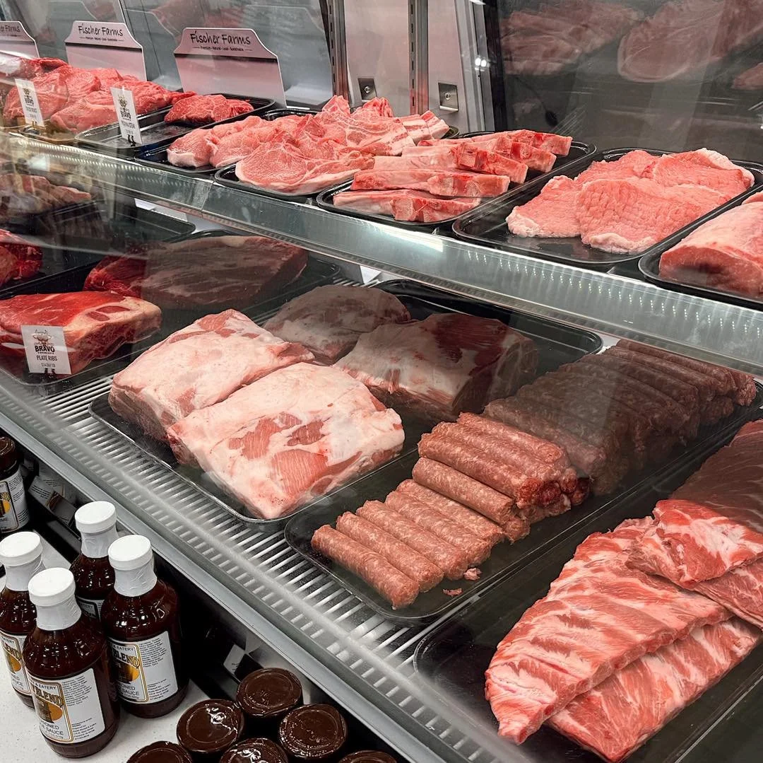 A meat display case from Bravo Butcher Shop at Roots Market with various cuts of beef, including steaks, ribs, ground beef, and sausages, in a grocery store.