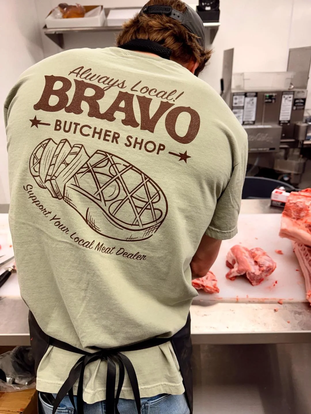A person wearing a green T-shirt with a graphic of a meat cleaver and text that reads, 'Always Local! BRAVO Butcher Shop'. The person is preparing meat in a butcher shop, with raw meat on a white cutting table in front of them.
