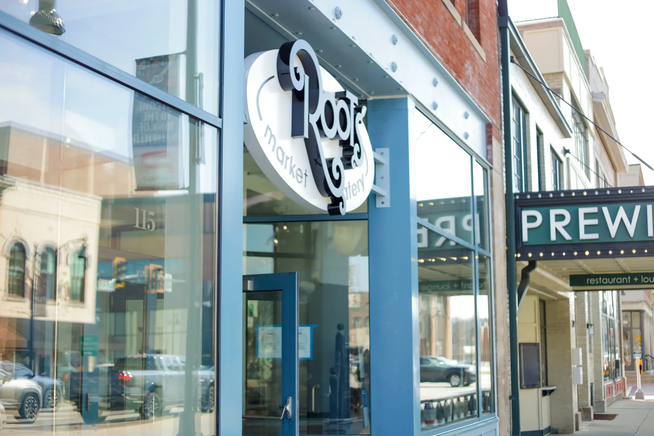 Exterior of Root Market with large glass windows, a sign reading 'Root Market,' and a partially visible sign for Prew.