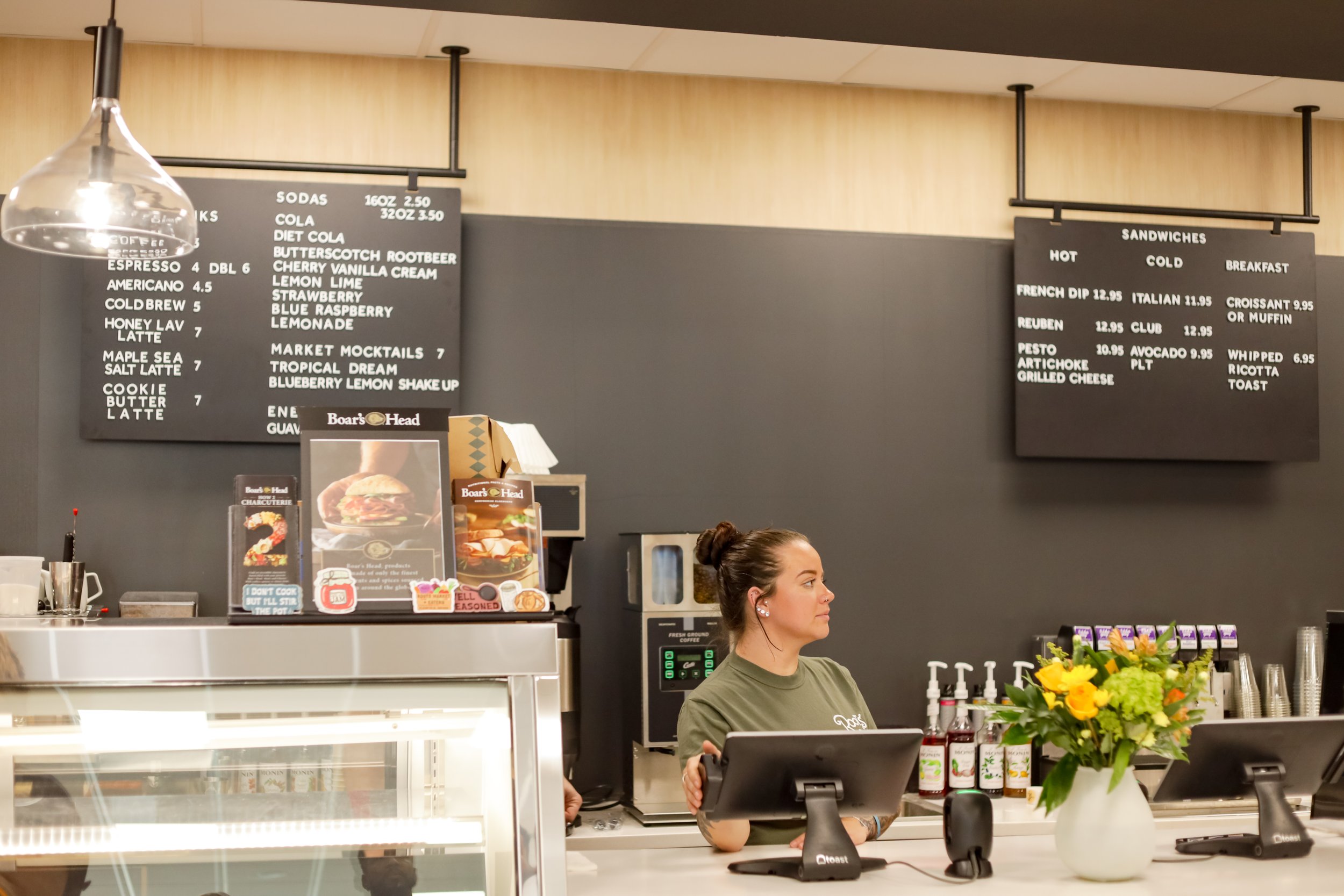 A woman at a café counter with a bouquet of flowers and two register screens, behind her are menu boards with drinks and sandwiches.