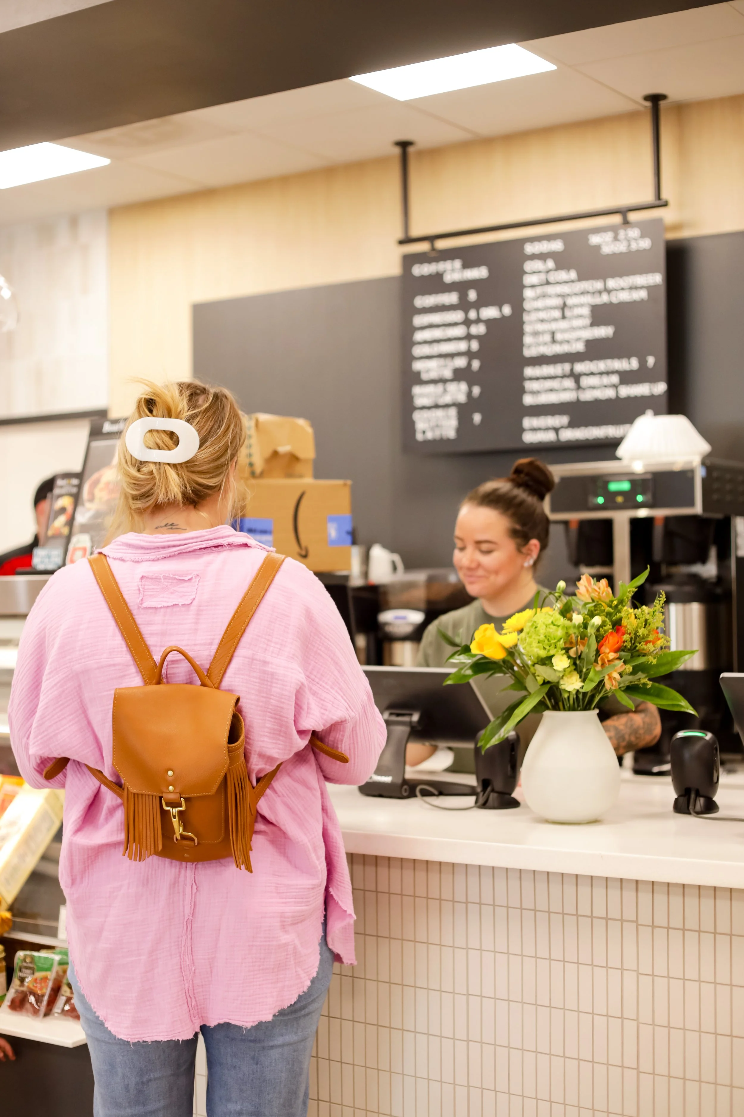 A woman at Roots Market with a pink shirt and a tan backpack at a cafe counter, ordering with a barista behind the counter, floral arrangement on the counter.