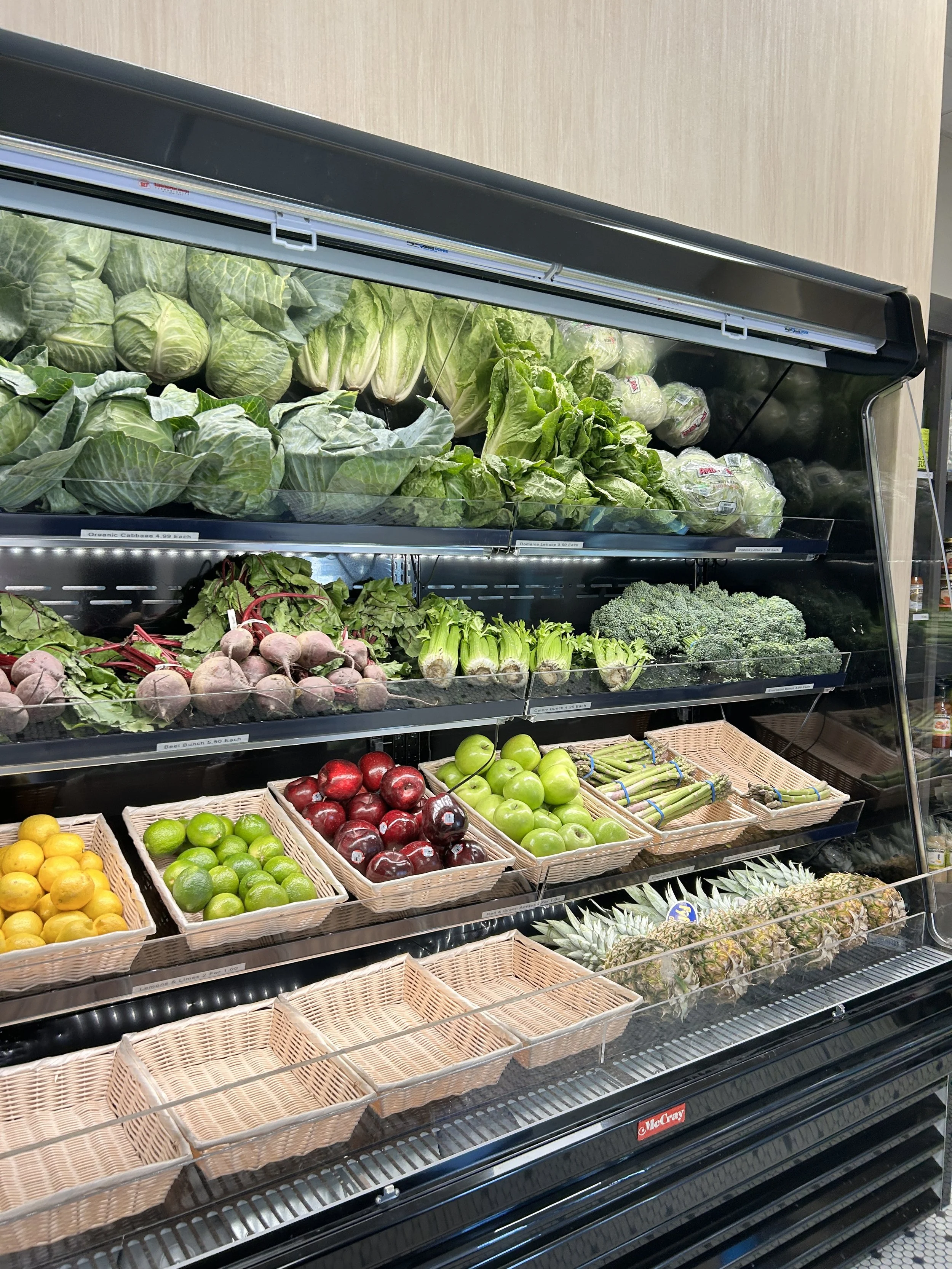 Refrigerated display case at Roots Market filled with fresh vegetables and fruits, including cabbage, lettuce, broccoli, radishes, celery, apples, lemons, limes, pineapples, and empty baskets.