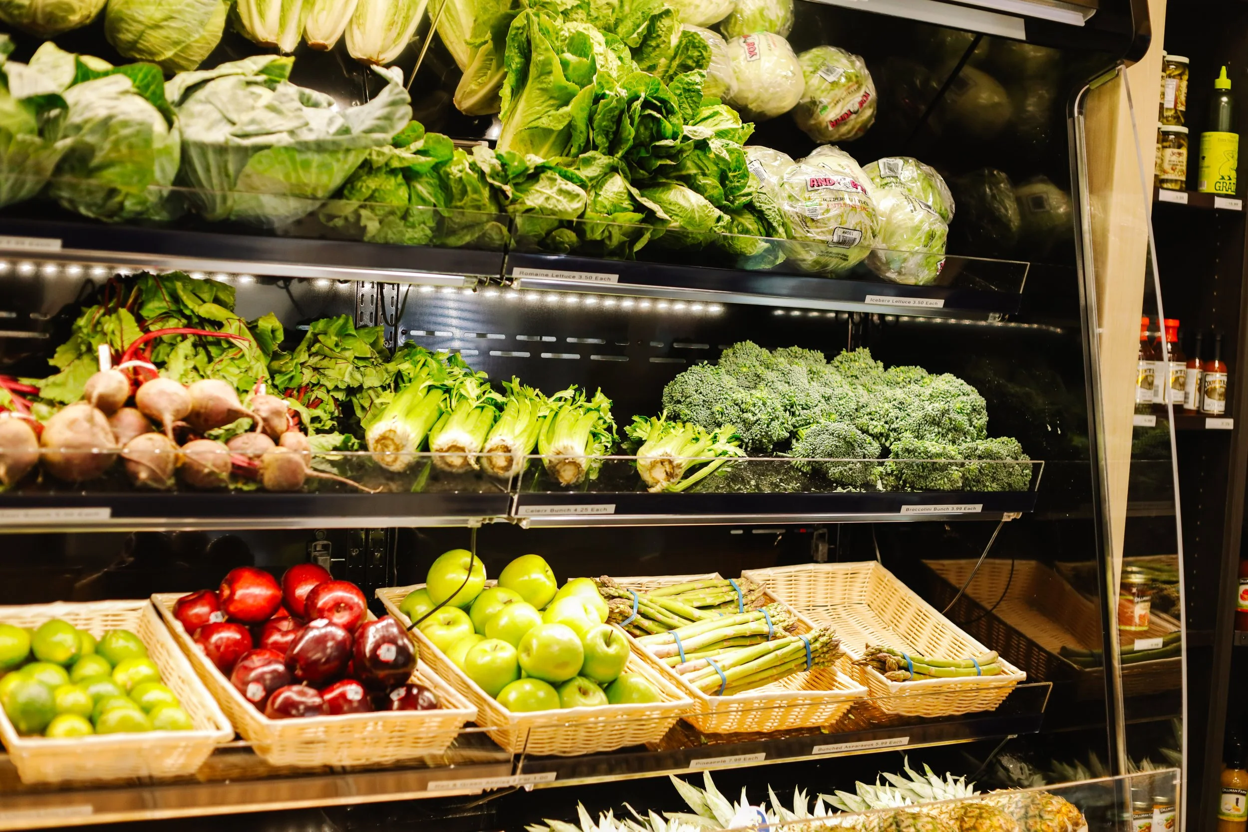 Fresh vegetables at Roots Market and Eatery on display in a grocery store, including lettuce, broccoli, asparagus, apples, and beets.