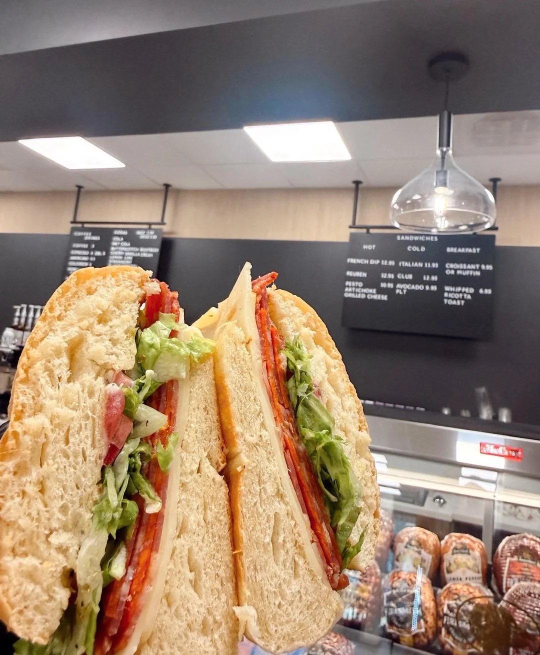 Close-up of a club sandwich with lettuce, tomato, cheese, turkey, and bacon on toasted bread, held in front of a Roots Market and Eatery menu board.