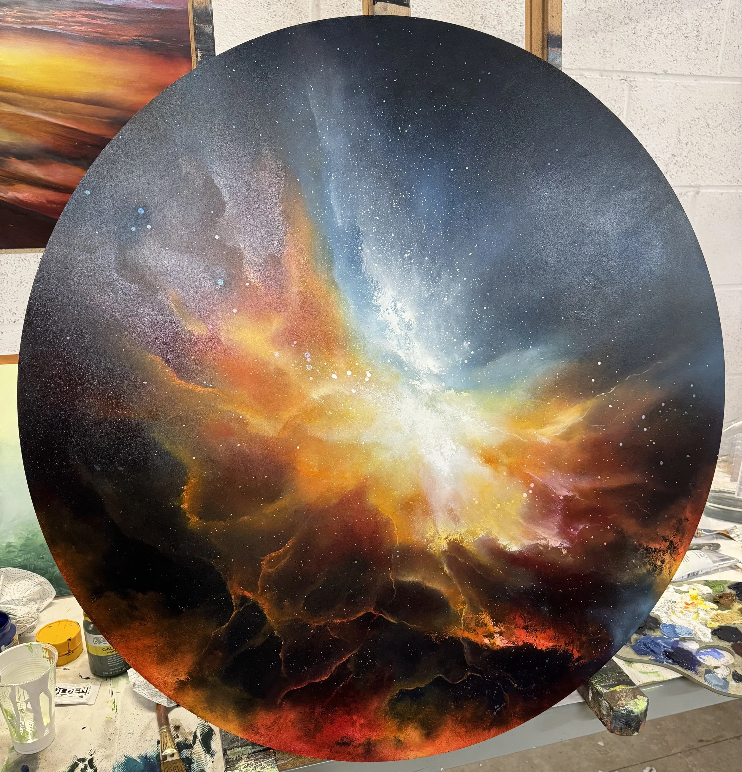 A round canvas painting of a vibrant galaxy scene with bright stars, swirling clouds of orange, yellow, blue, and red cosmic dust, and a dark space background.