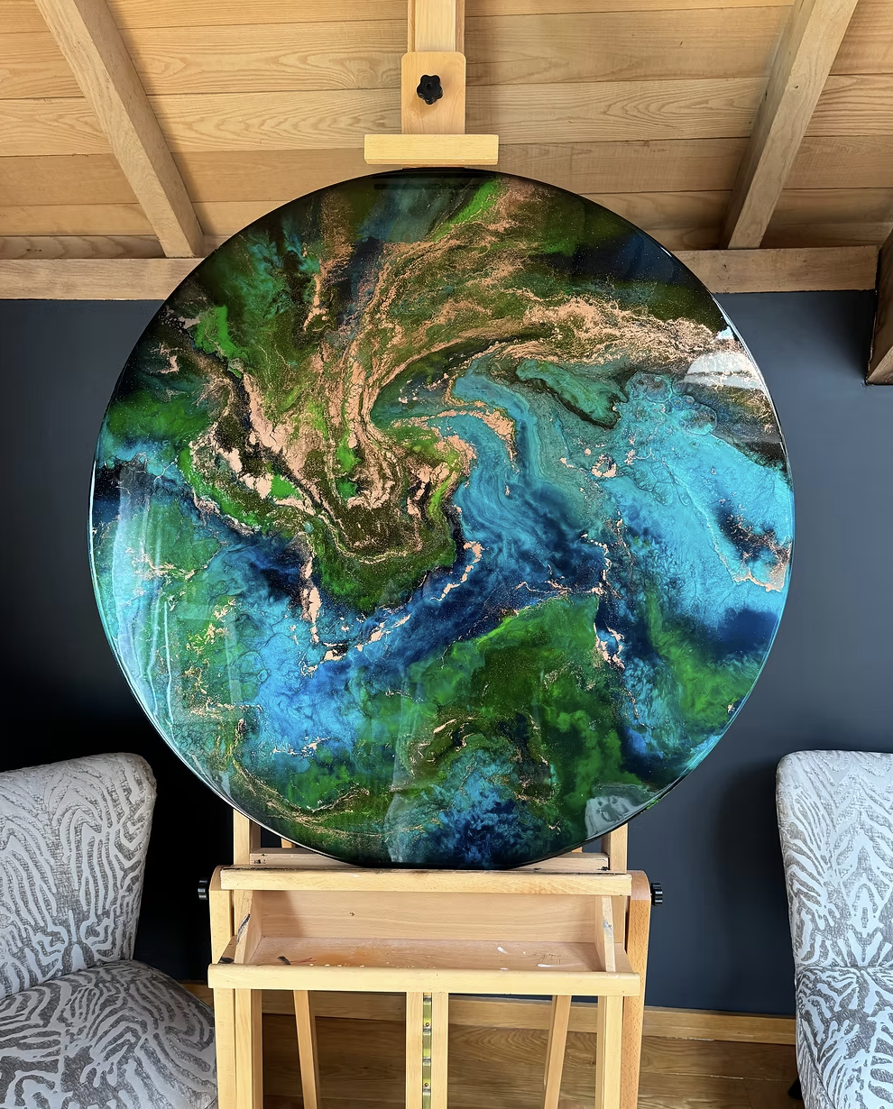 Circular abstract painting with green, blue, black, and gold colors on an easel in a room with wood ceiling and two patterned chairs.