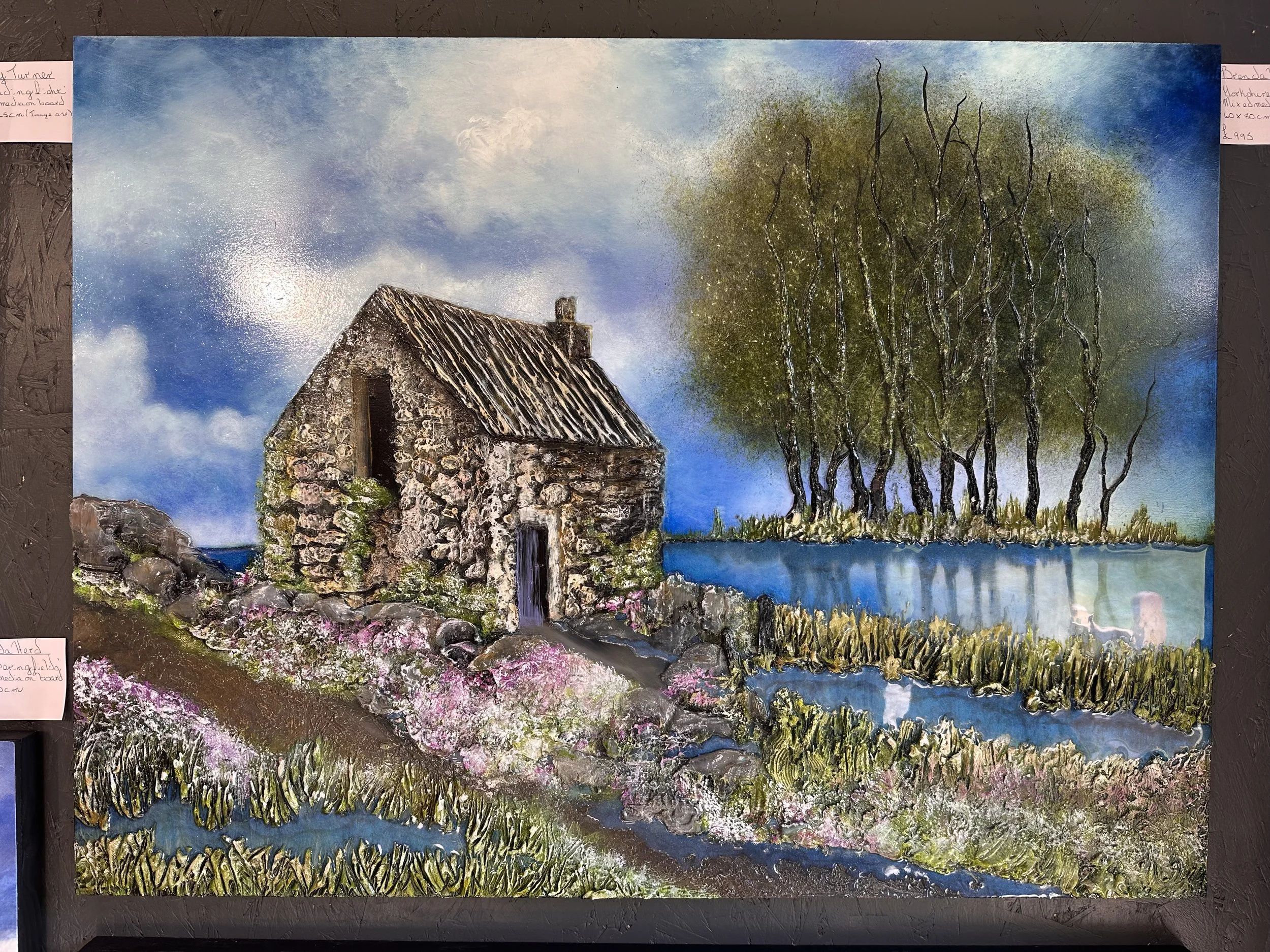 A painting of a rustic stone house beside a body of water, with a large green tree and a blue sky with clouds in the background.