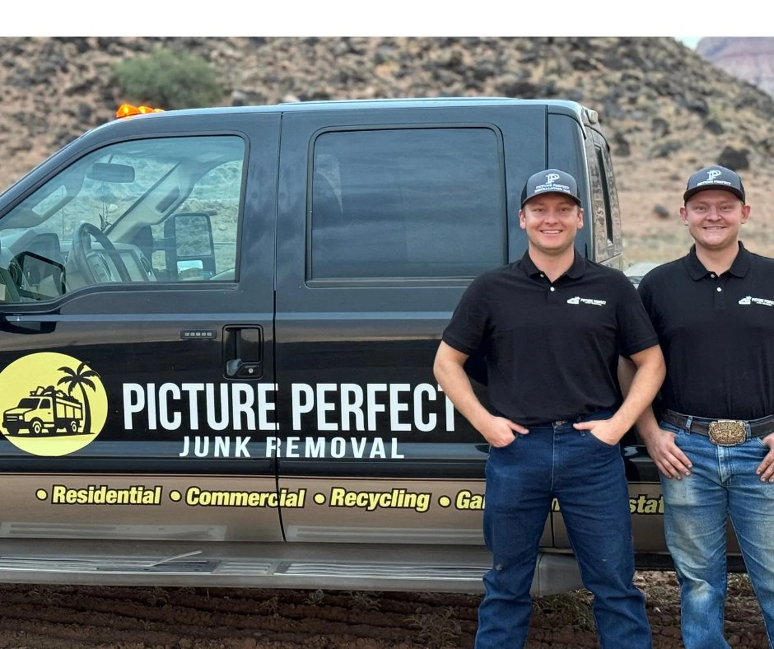 Two men standing in front of a black truck with the logo and name 'Picture Perfect Junk Removal' on the side. The truck has a yellow circle logo with a palm tree and a van. The men are wearing black collared shirts and caps, smiling, with a desert or