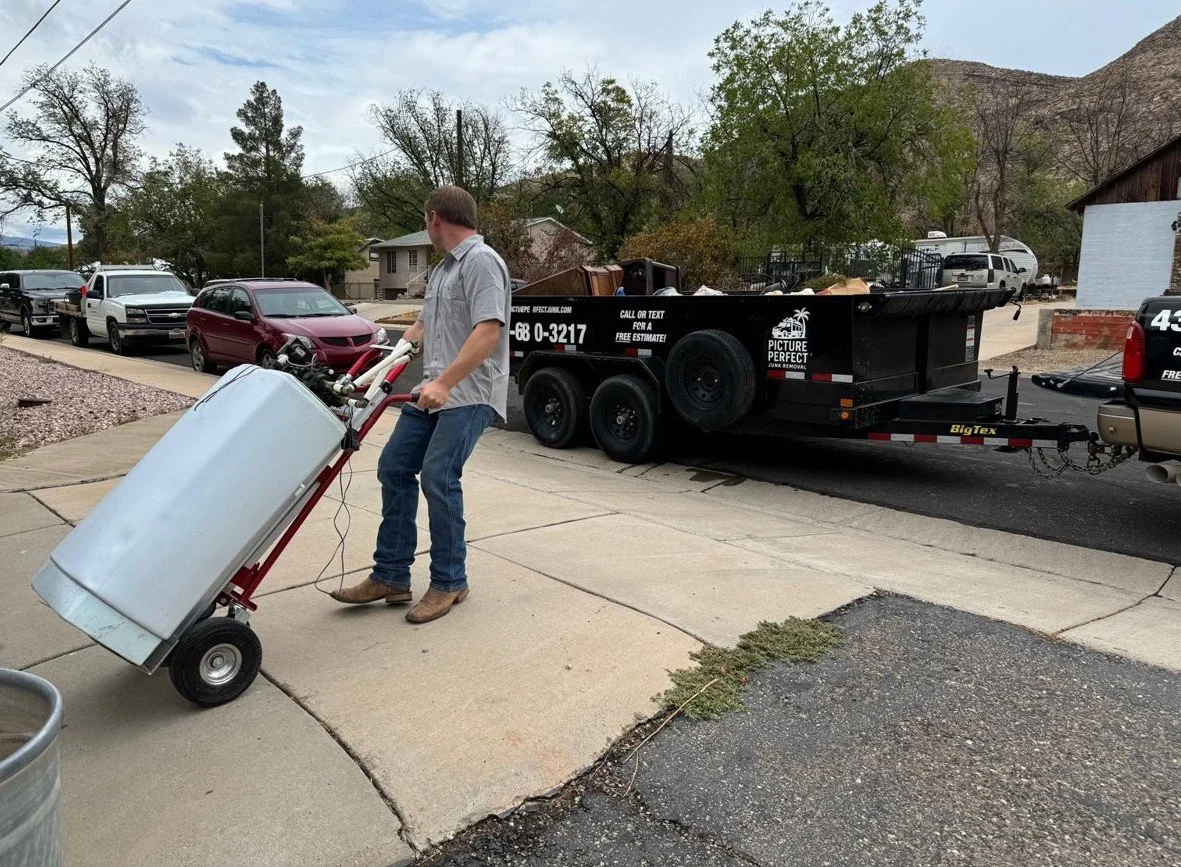 Man moving a metal appliance on a hand truck toward a black trailer parked on the street, in a residential neighborhood with trees and mountains in the background.
