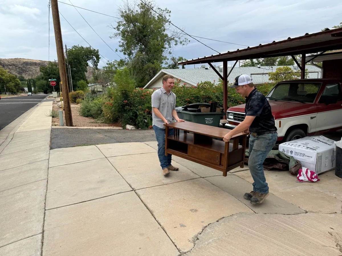 Two young men are moving a wooden piece of furniture outside, one on each side, smiling at each other. They are on a sidewalk in a suburban neighborhood with a carport, a red and white truck, and boxes nearby.