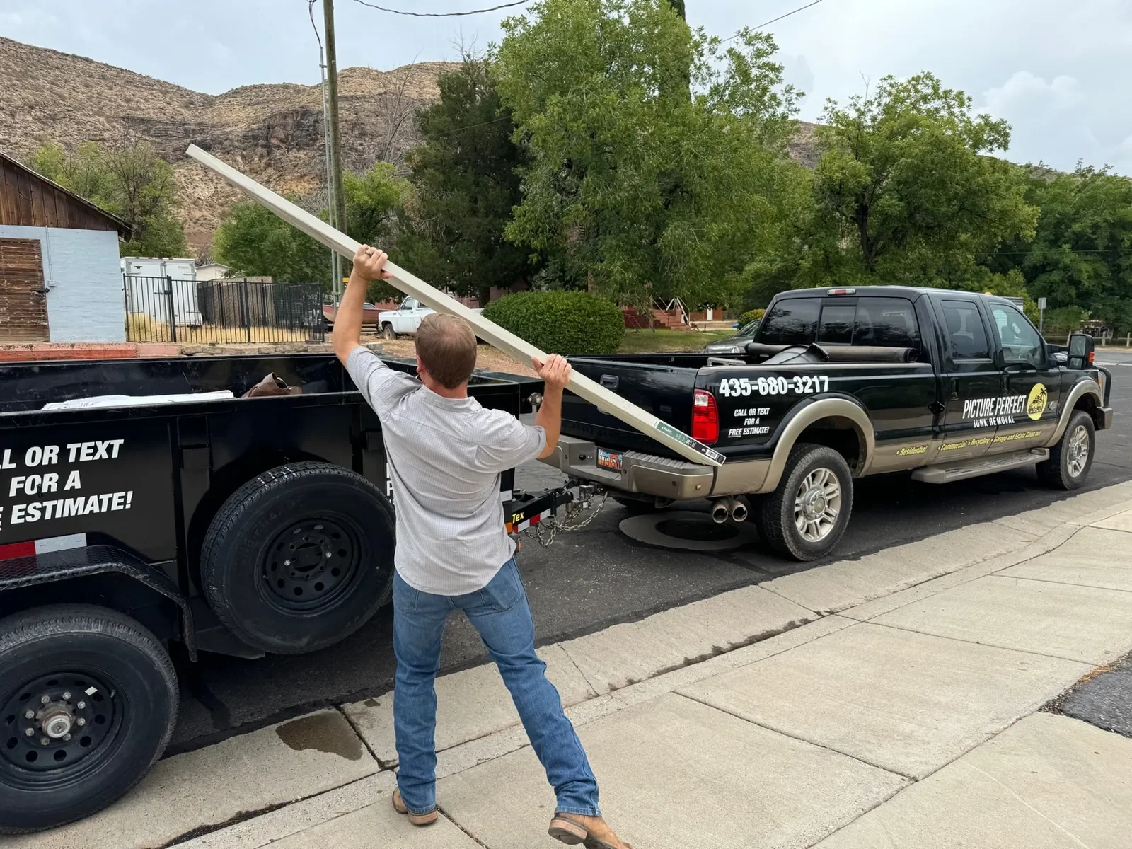 A man is putting a piece of an awning or similar item into the bed of a black pickup truck on the street. The truck has business branding and contact information, and is parked next to a sidewalk with houses and trees in the background.
