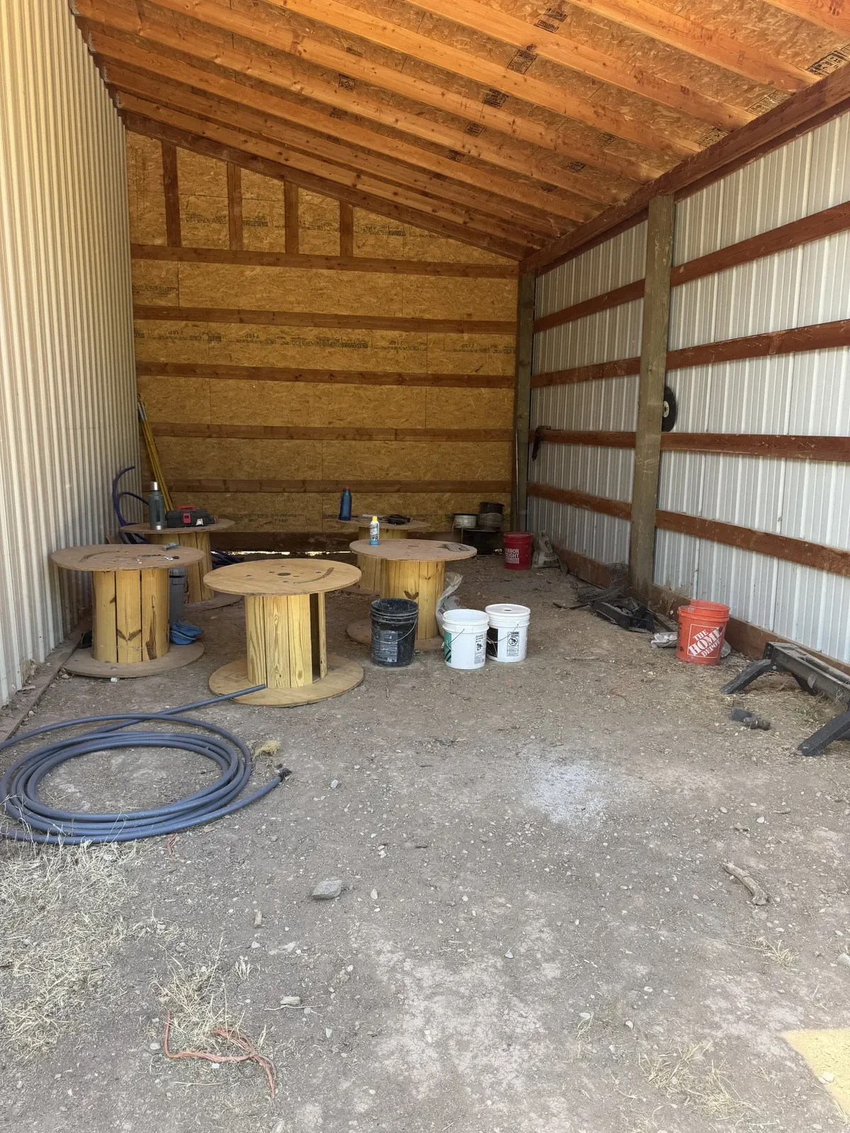 Interior of a partially finished barn or shed with a dirt floor, woodenworktables, buckets, coiled hoses, and construction tools along the walls.