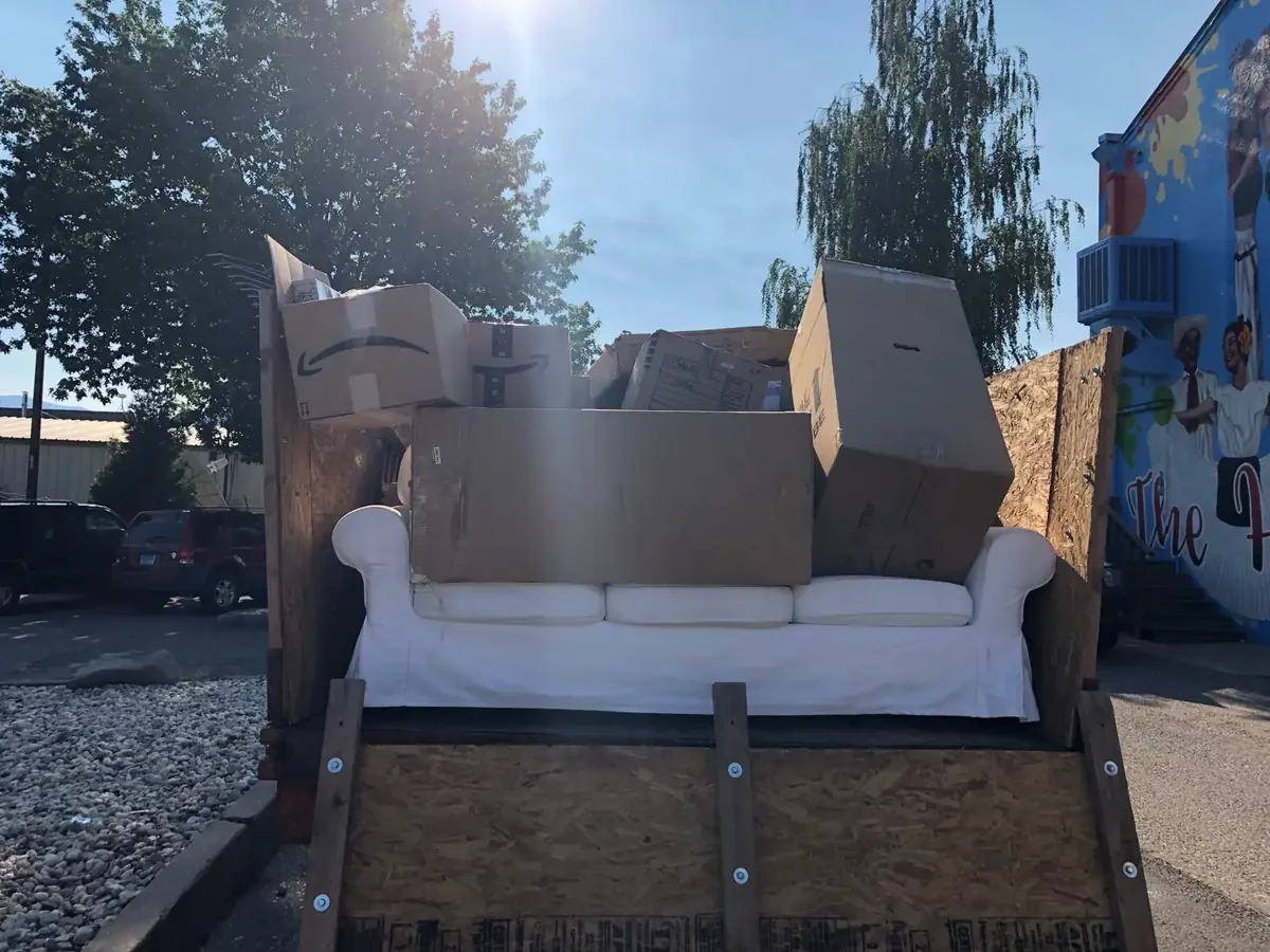 A white sofa with boxes on top, set up on a platform outdoors, with parking cars and trees in the background.