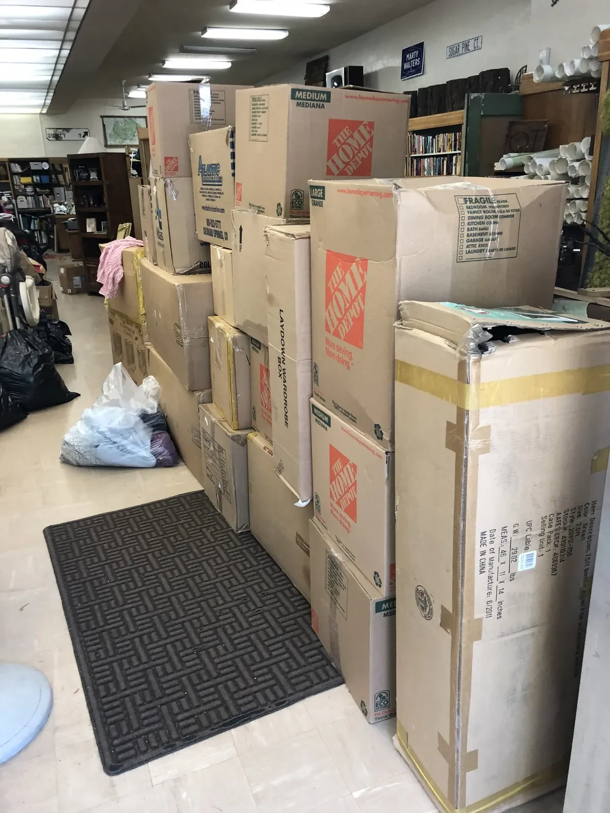 Several packed moving boxes stacked in a store aisle with a black rug on the floor, surrounded by miscellaneous items and shelves.