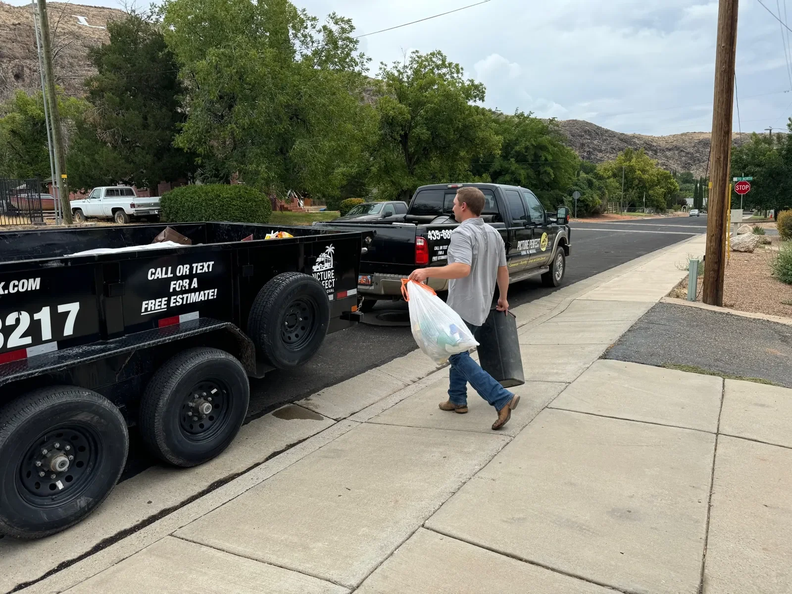 A man is walking on the sidewalk carrying a large plastic bag filled with trash in one hand and a trash can in the other, heading towards a black trash hauling truck parked nearby.