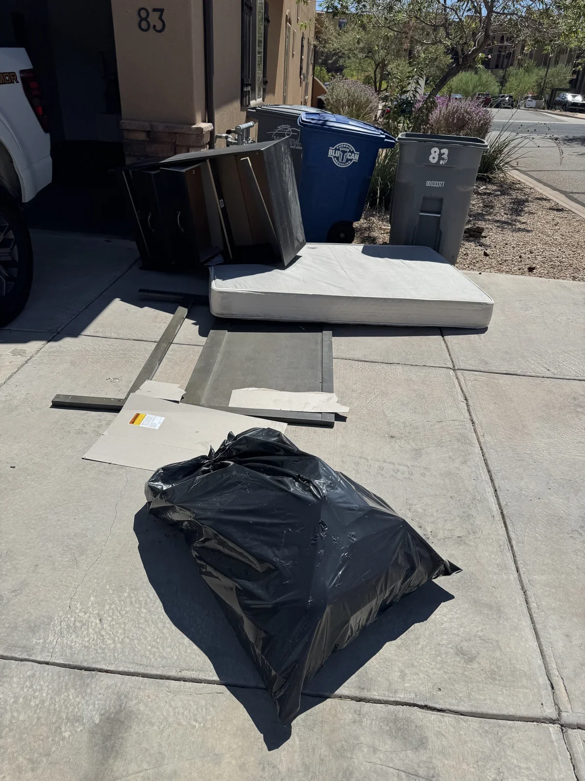 Disassembled furniture, mattress, and trash bags on sidewalk near trash bins and building, with trees and parked cars in background.