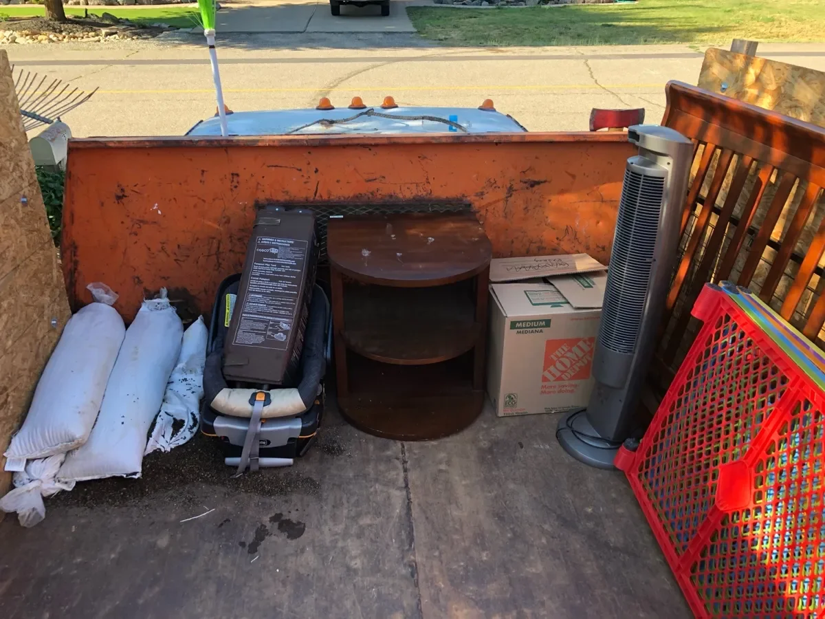 Miscellaneous household items including sandbags, a car seat, a small wooden table, a boxed fan, a boxed chair, a tall tower fan, and a red plastic crate on a concrete surface outside.