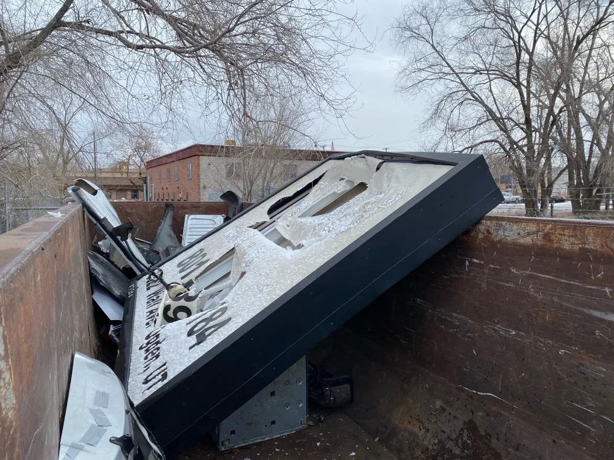 A destroyed and overturned large electronic sign or billboard inside a rusty container or dumpster, with debris scattered around and leafless trees in the background.