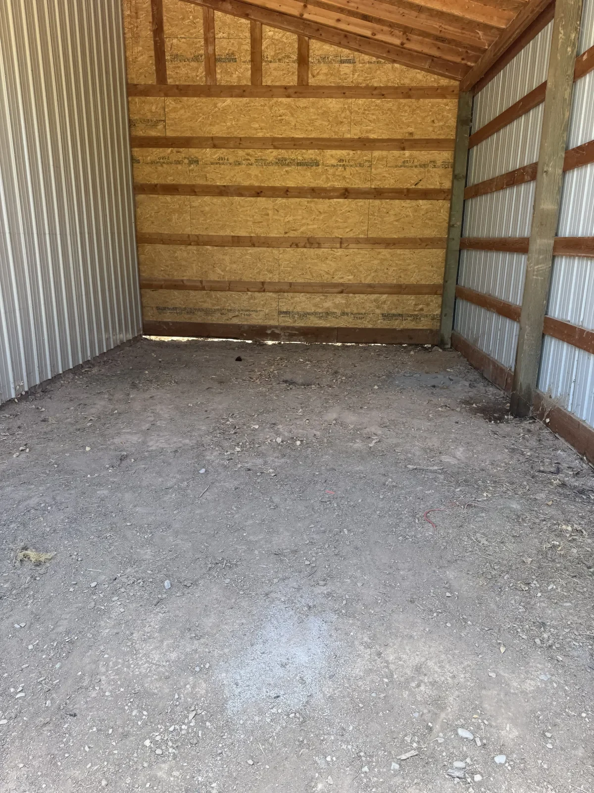 Interior of a small, unfinished garage or storage shed with bare dirt floor, metal siding on one wall, and wooden framing with OSB panels on the back wall and ceiling.