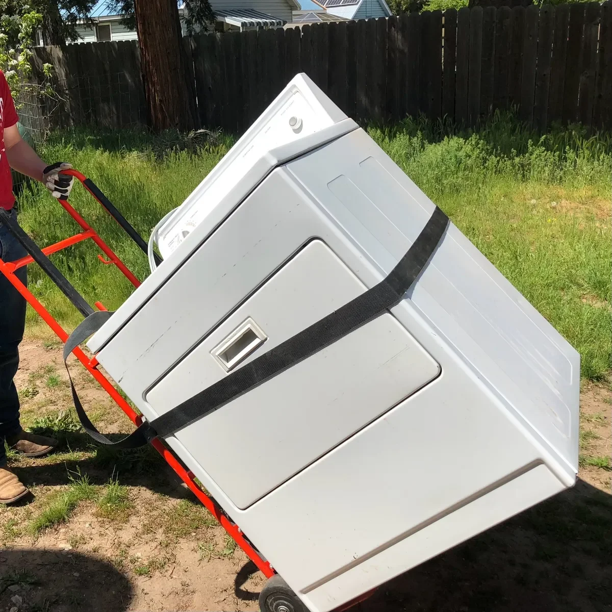 A person is using a hand truck to move a white washing machine outdoors on grass and dirt, with a wooden fence and some houses in the background.