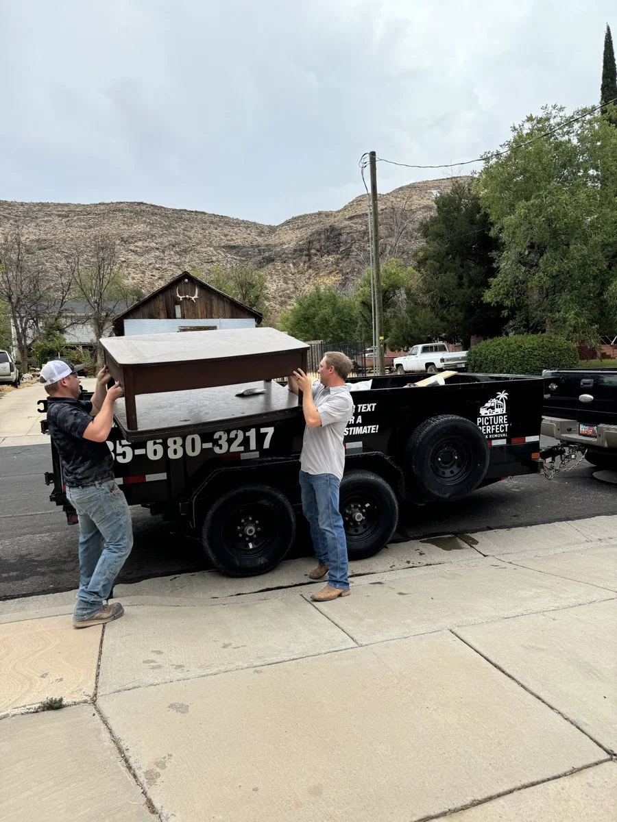 Two men are loading a small wooden table onto a black trailer parked on a street, with a mountain, trees, and some houses in the background.