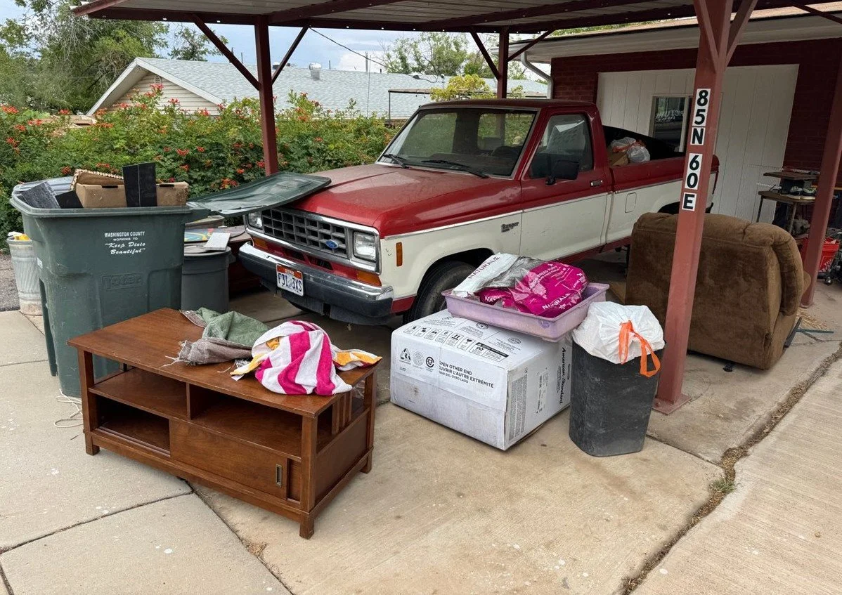 A red and white pickup truck parked under a carport next to house with various household items, including a brown couch, a wooden TV stand, a trash bin, a plastic storage container, and boxes, cluttered around it.