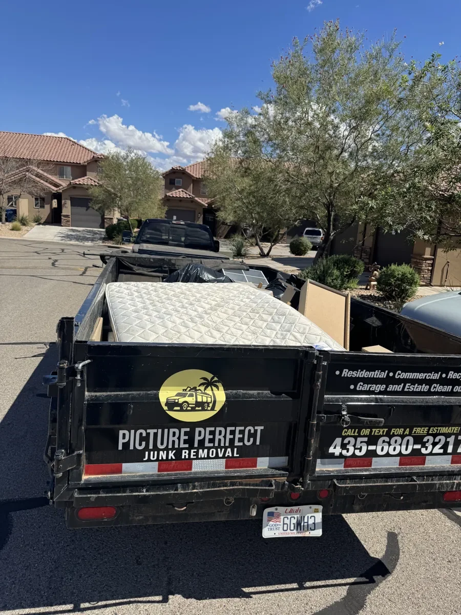 A black trailer with the logo and contact information of 'Picture Perfect Junk Removal' is parked in a neighborhood. It is loaded with a mattress, furniture, and other household items, indicating junk removal activity.