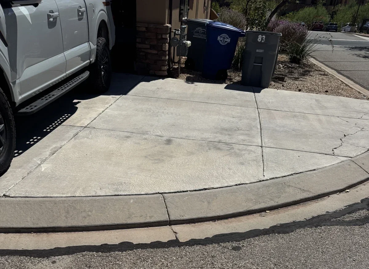 Concrete sidewalk and driveway with a parked silver truck and two trash bins near a brick wall, street in the background.