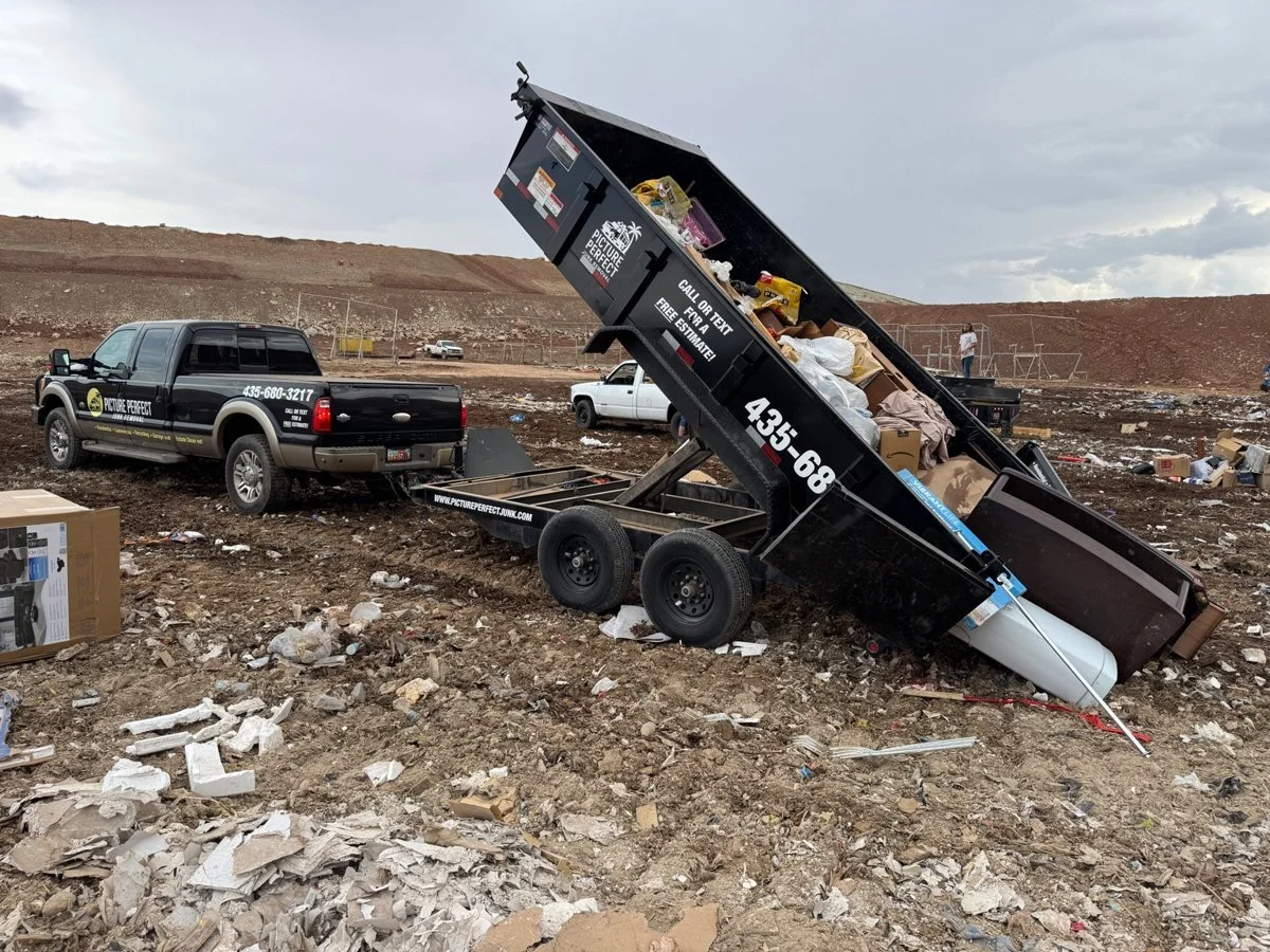 A dump truck loaded with trash and debris, tipped on the ground, attached to a pickup truck at a landfill or junkyard with more scattered trash and a hill in the background.