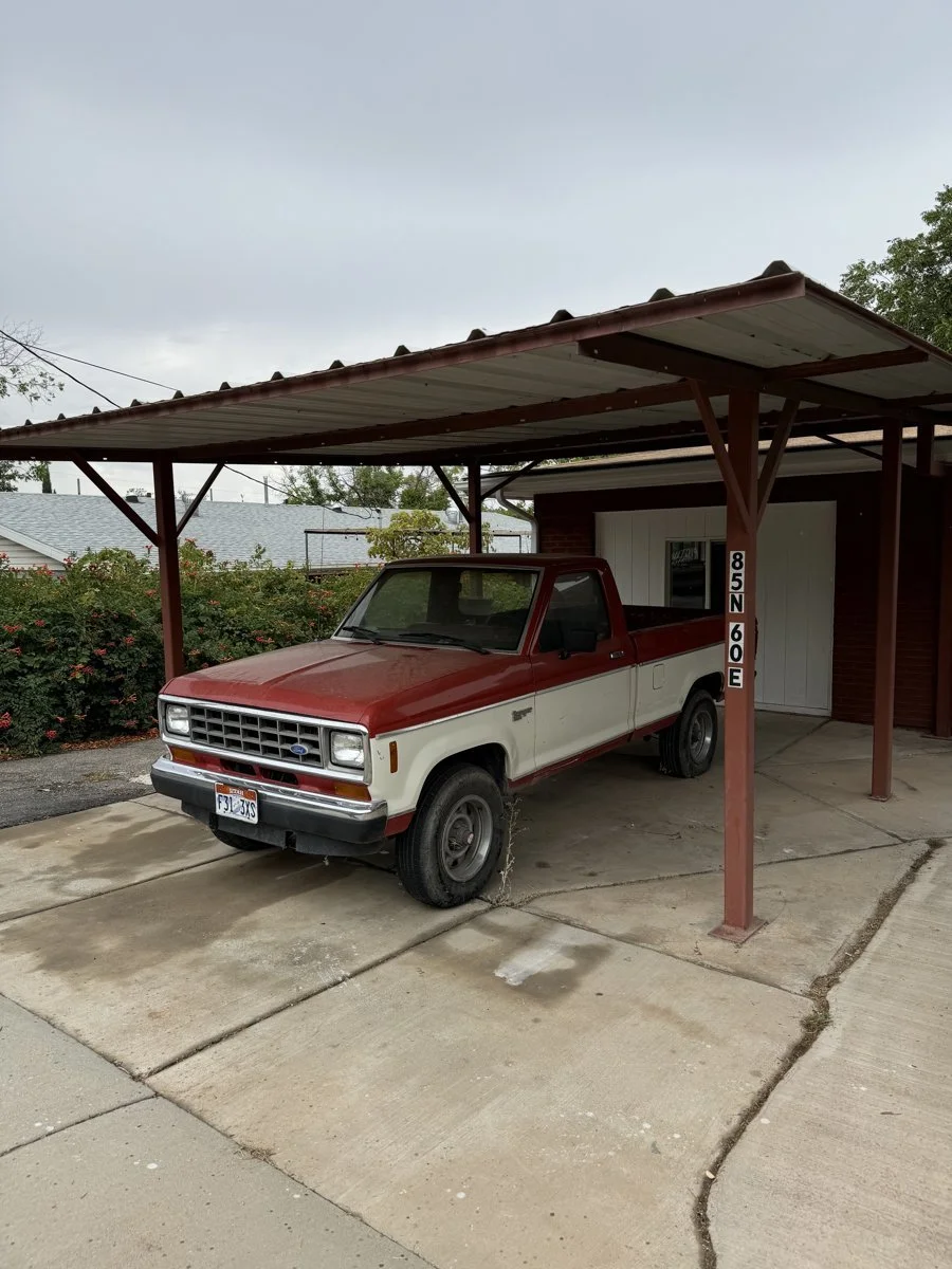 Old red and white pickup truck parked under a carport with a concrete driveway in front of a house.