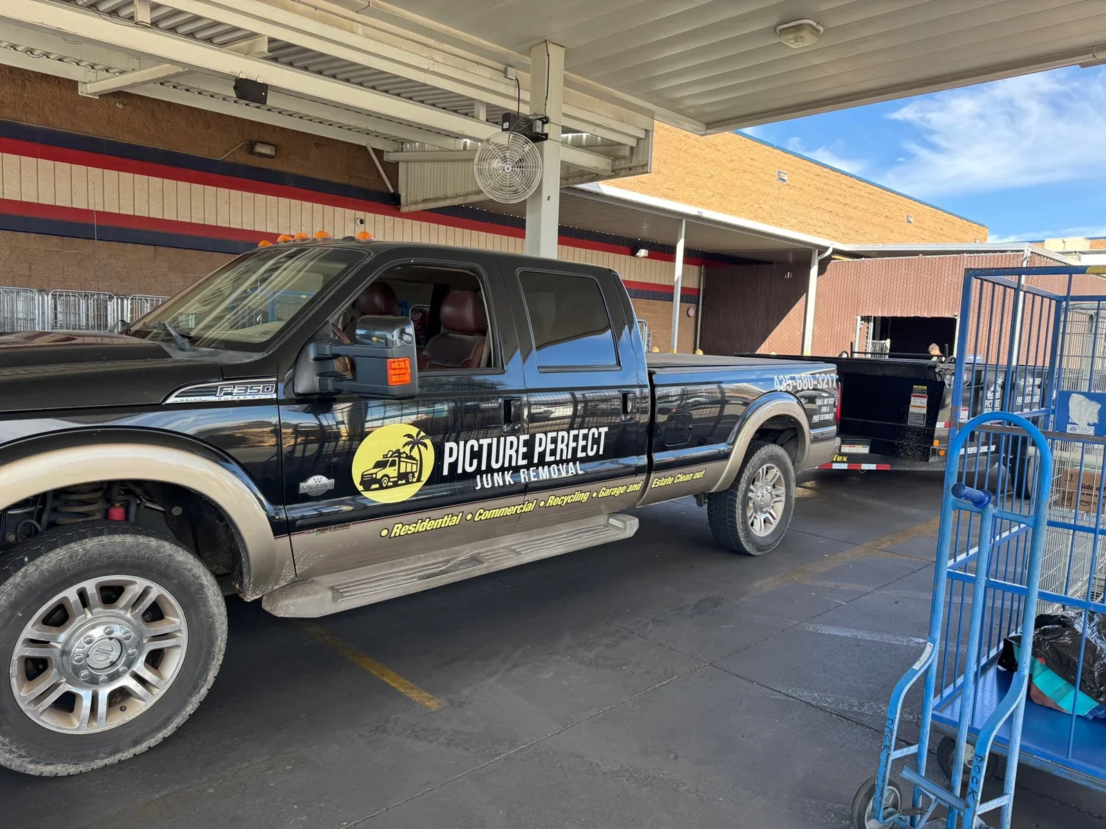Black pickup truck with company logo and text, parked at a vehicle removal service. The truck is towing a black trailer. The setting appears to be a parking lot or service area with shopping carts on the right and a building in the background.