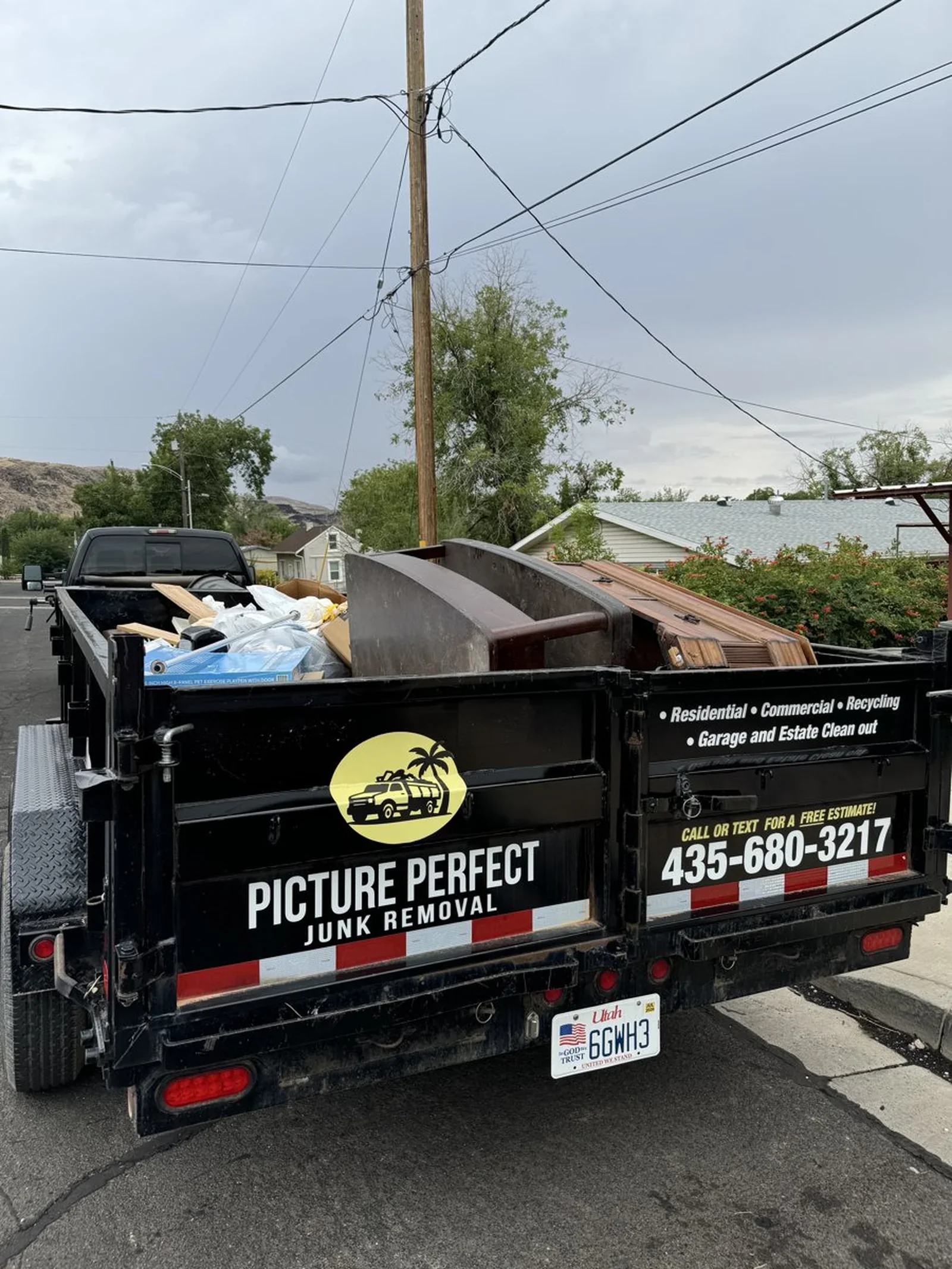 A black junk removal truck with the company name "Picture Perfect Junk Removal" and a phone number parked on the street, loaded with various items like wood and boxes, under a cloudy sky.