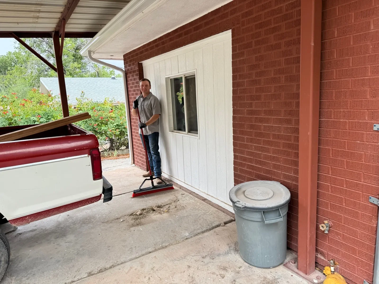 A man in a gray shirt and jeans holding a rake and a garden tool standing on a patio under a carport, smiling at the camera with a pickup truck nearby and greenery in the background.