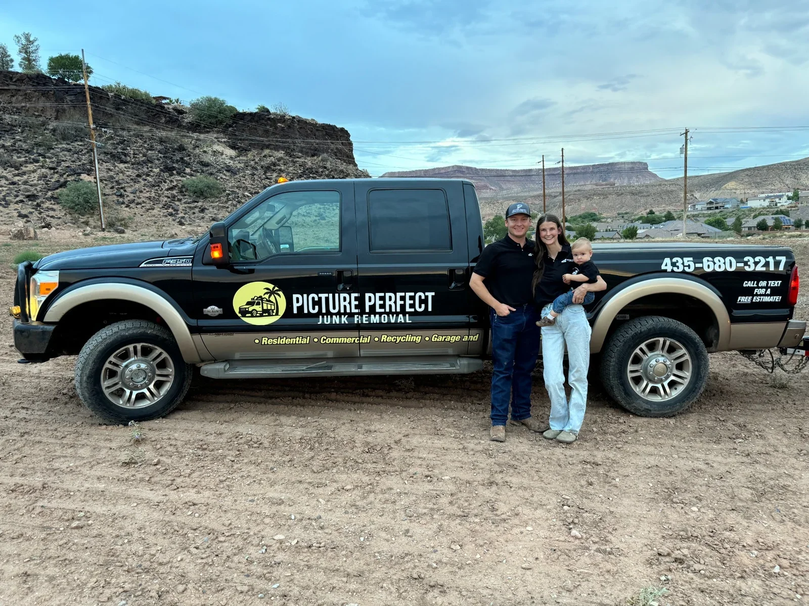 A family of three standing together in front of a black Picture Perfect Junk Removal truck outdoors. The man, woman, and young child are smiling, with the child in the woman's arms. The truck displays contact information and services offered.