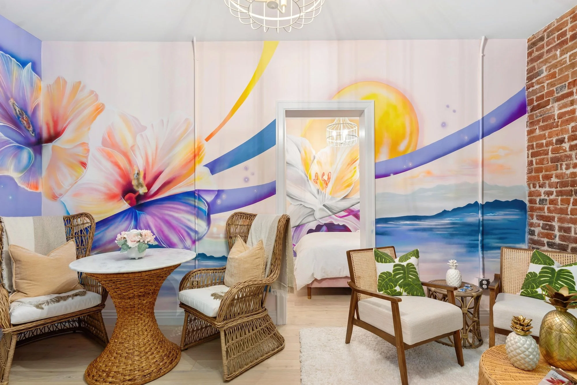 A colorful room with a large mural of flowers, a lake, and a sun on the wall. The room has woven furniture, a small round table with pink flowers, and green-leaf patterned pillows on chairs. There is a bed visible through a doorway in the background, and a brick wall on the right side.