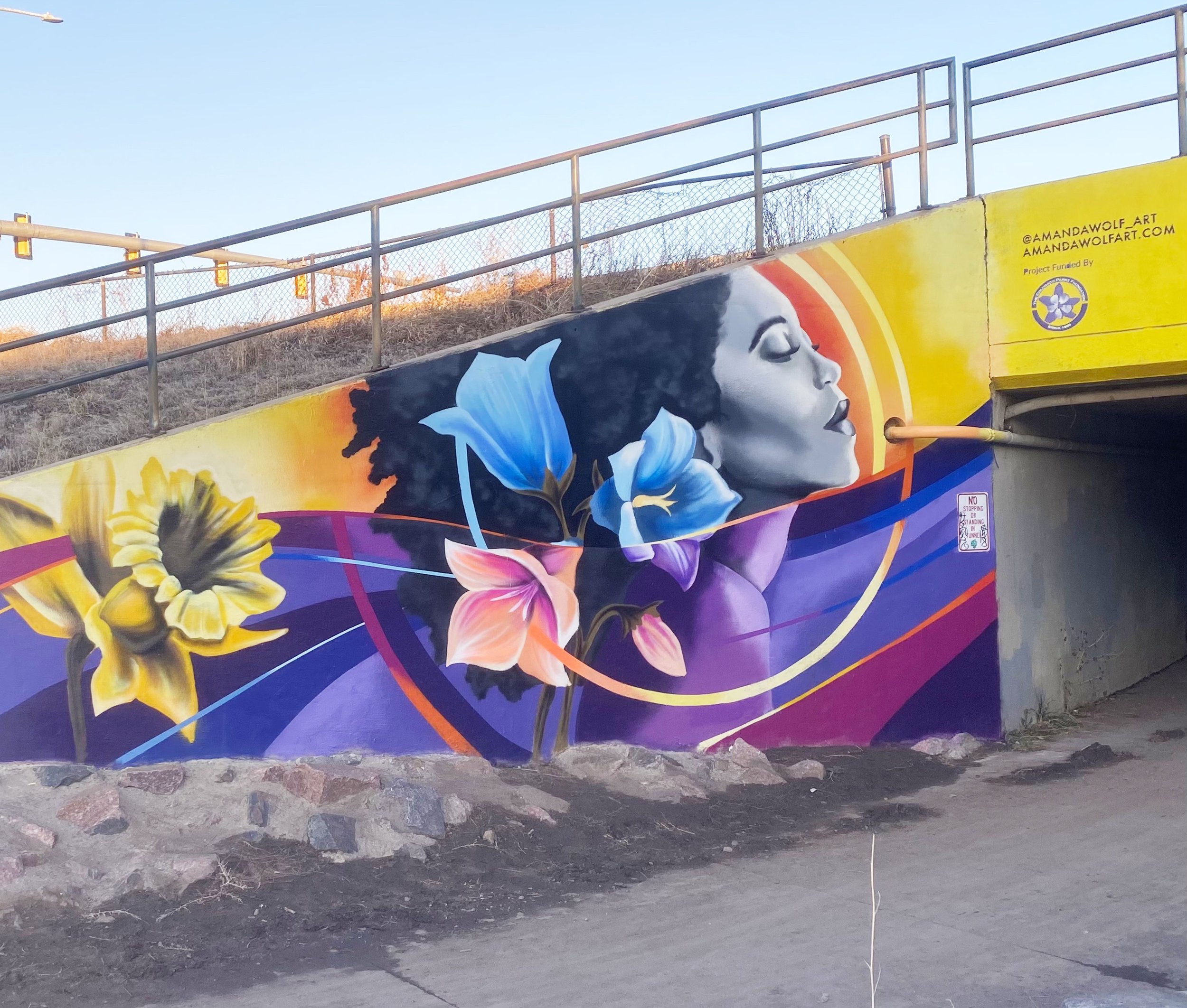 Colorful mural featuring a woman's profile with closed eyes and flowing hair, surrounded by vibrant flowers and abstract shapes, located under a bridge or tunnel.