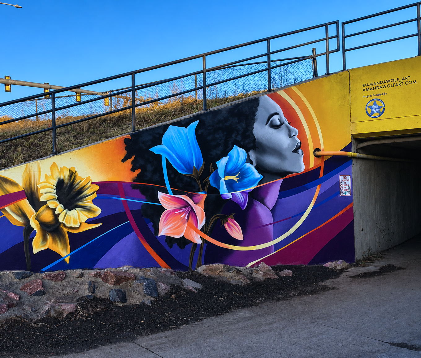 Colorful mural featuring a woman's profile with closed eyes and flowing hair, surrounded by vibrant flowers and abstract shapes, located under a bridge or tunnel.