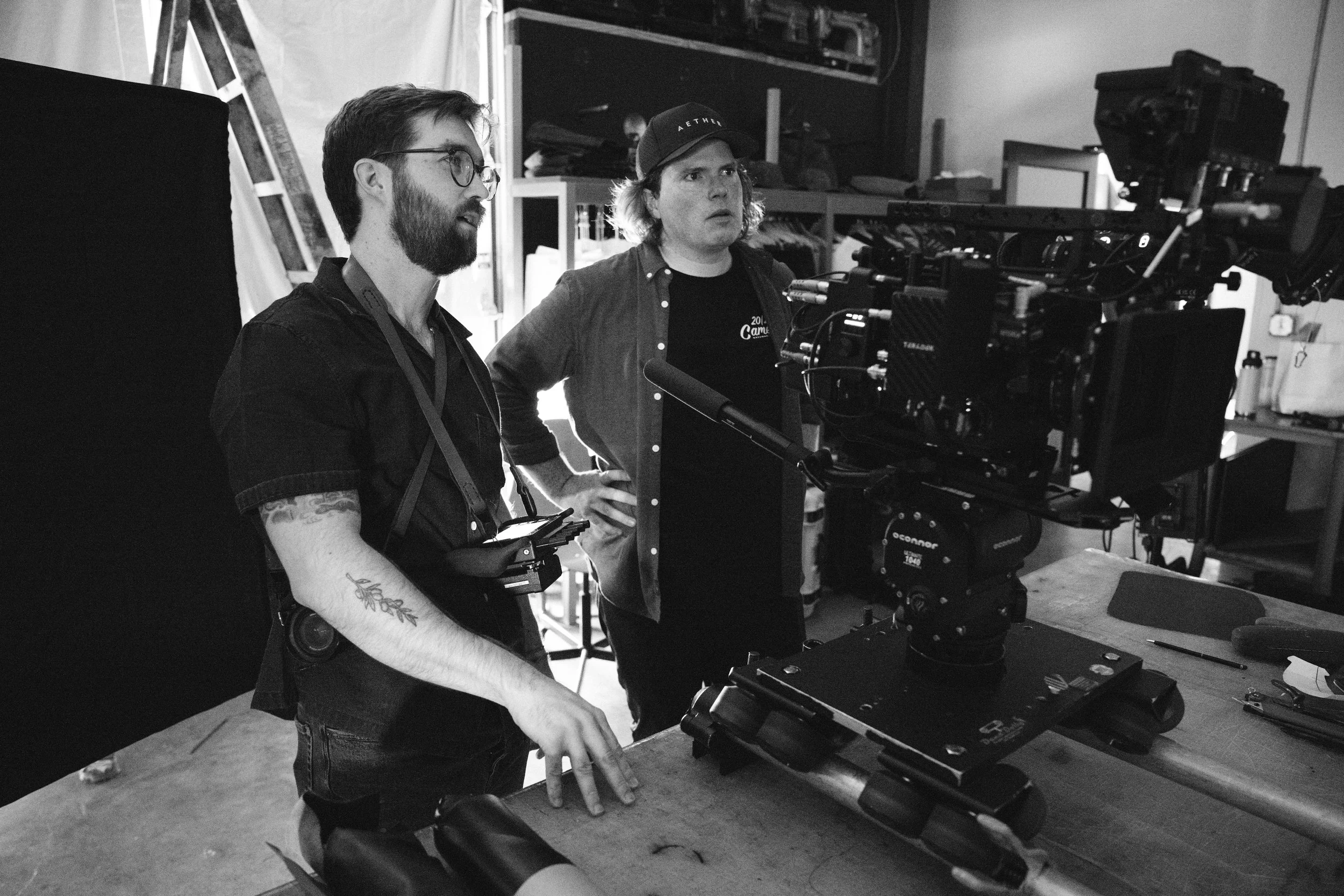 Two men operating a professional camera on a film set, with equipment and shelves in the background.