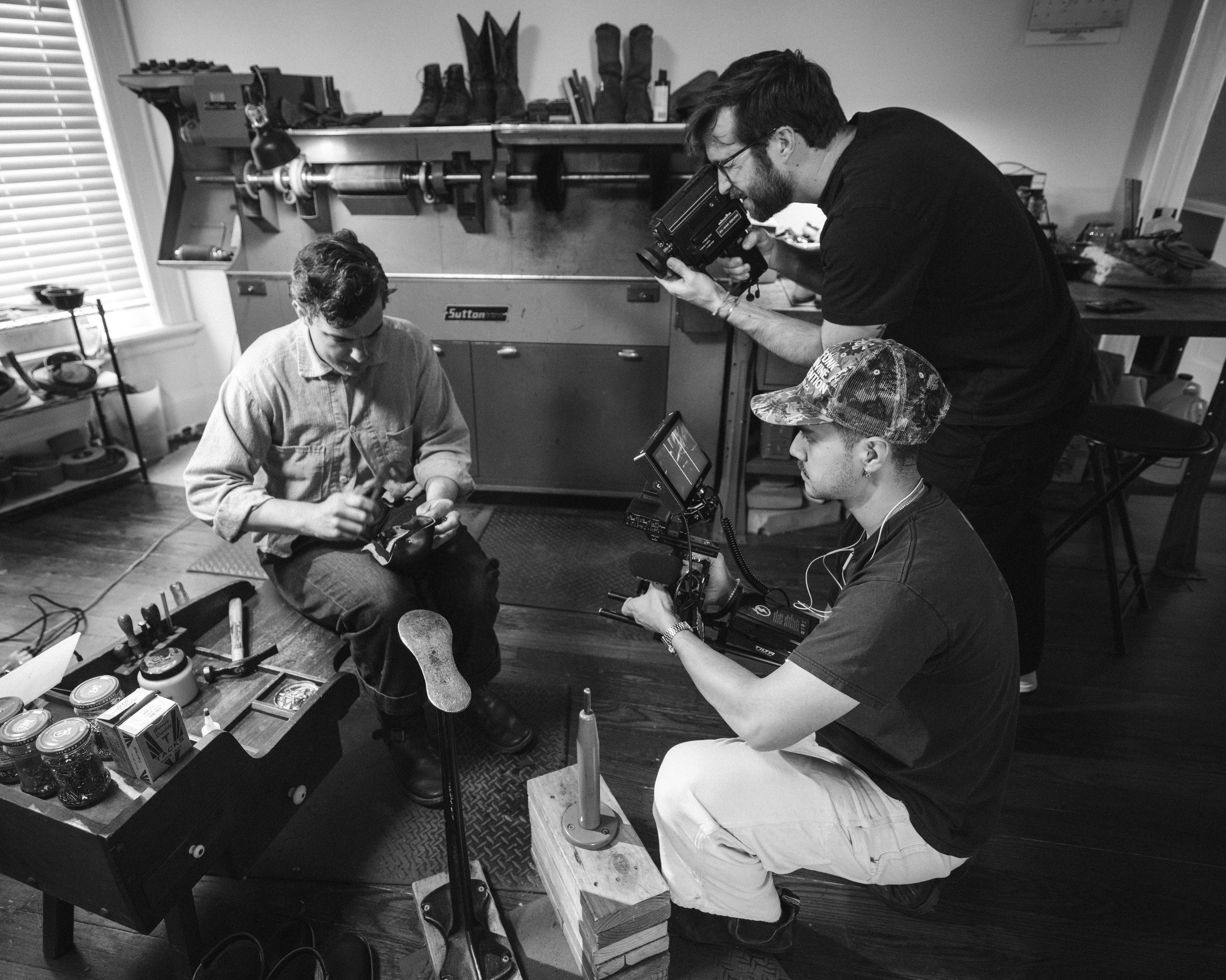 Three men filming a person in a workshop with tools and supplies around, using professional video equipment.