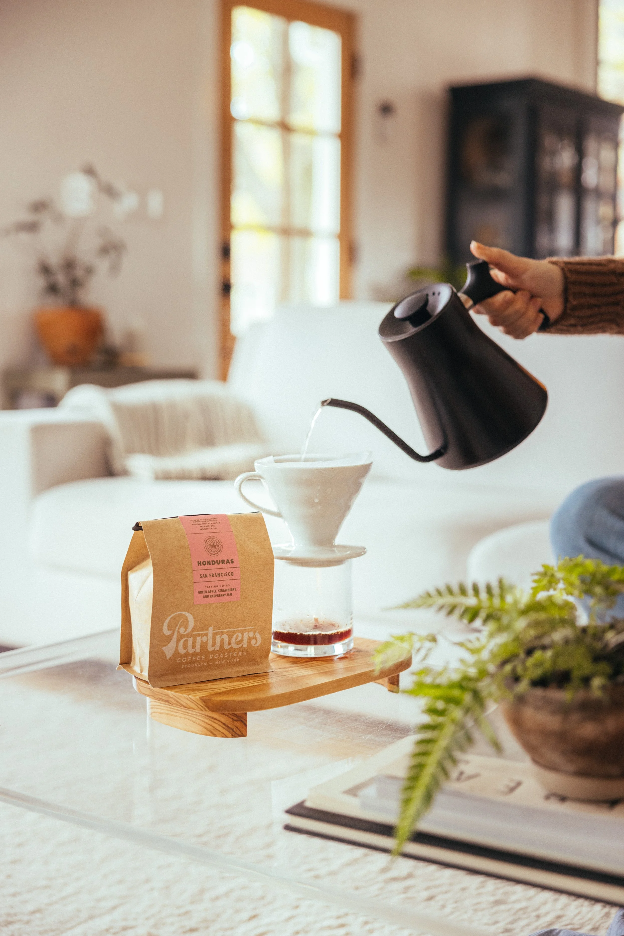A person pours hot water from a black gooseneck kettle into a white coffee dripper placed on a glass cup, with a bag of Partners Coffee Honduras coffee in the foreground, on a wooden tray on a coffee table in a cozy living room.