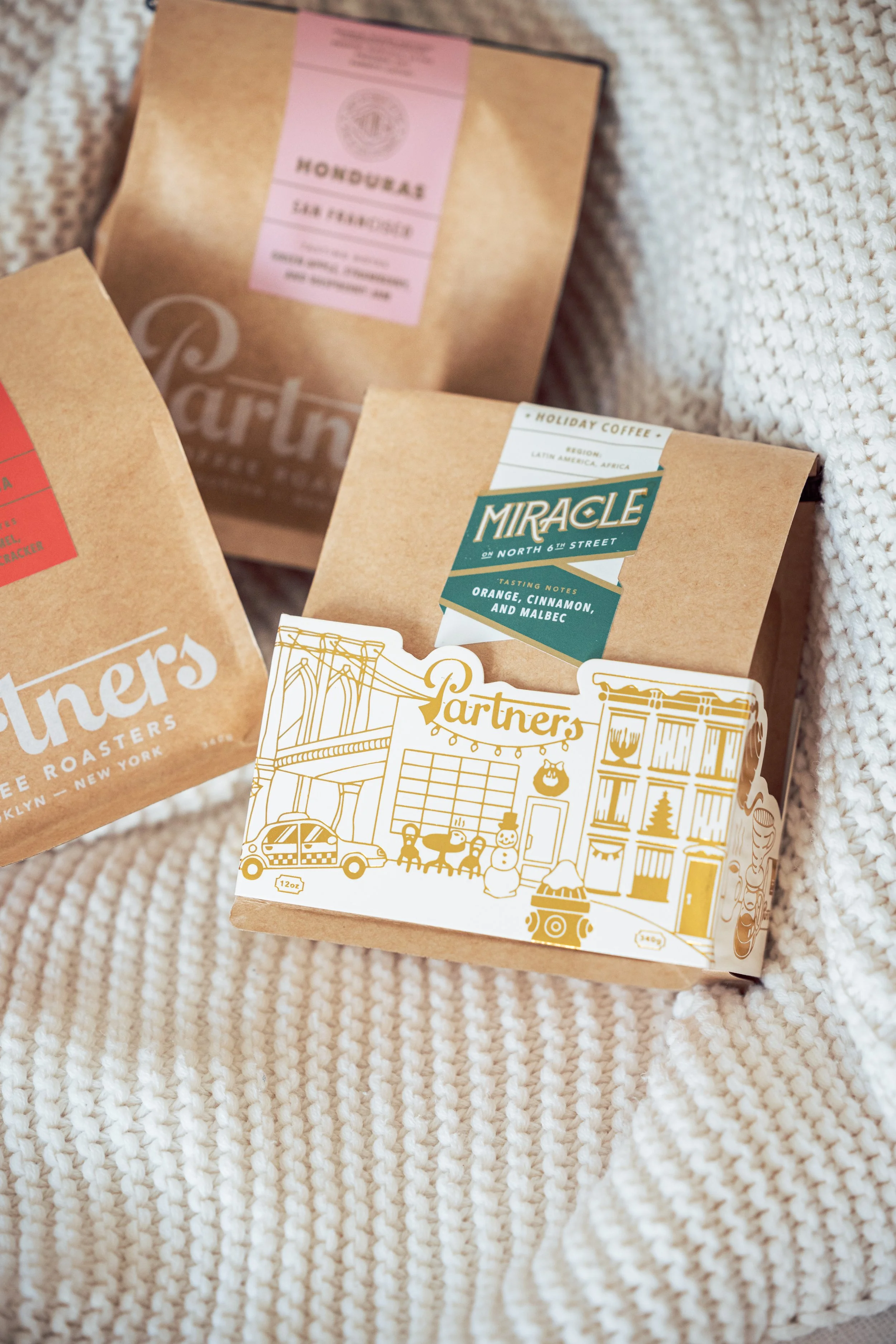 Commercial product photography Campaign for major coffee roaster Three packages of coffee resting on a knitted white blanket, with colorful labels indicating different flavors and origins.