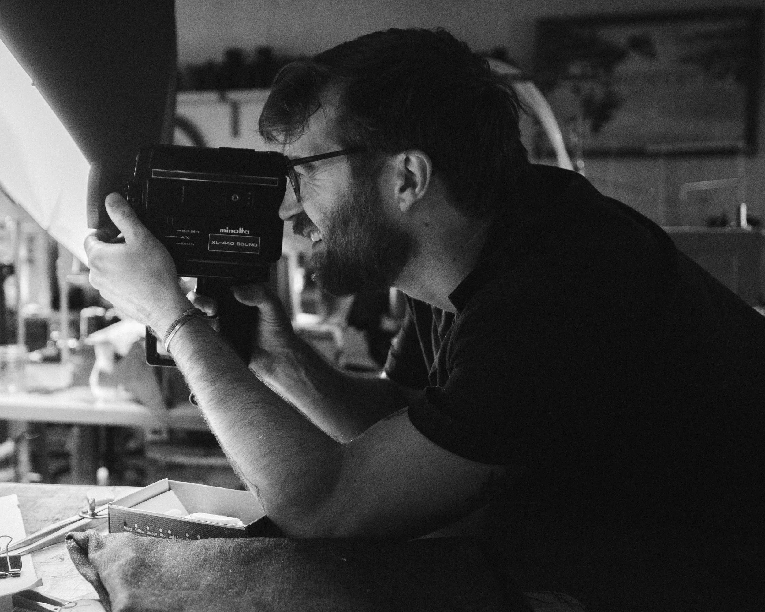 A man with glasses and a beard is looking through a vintage camera, smiling, in a workshop or studio setting.