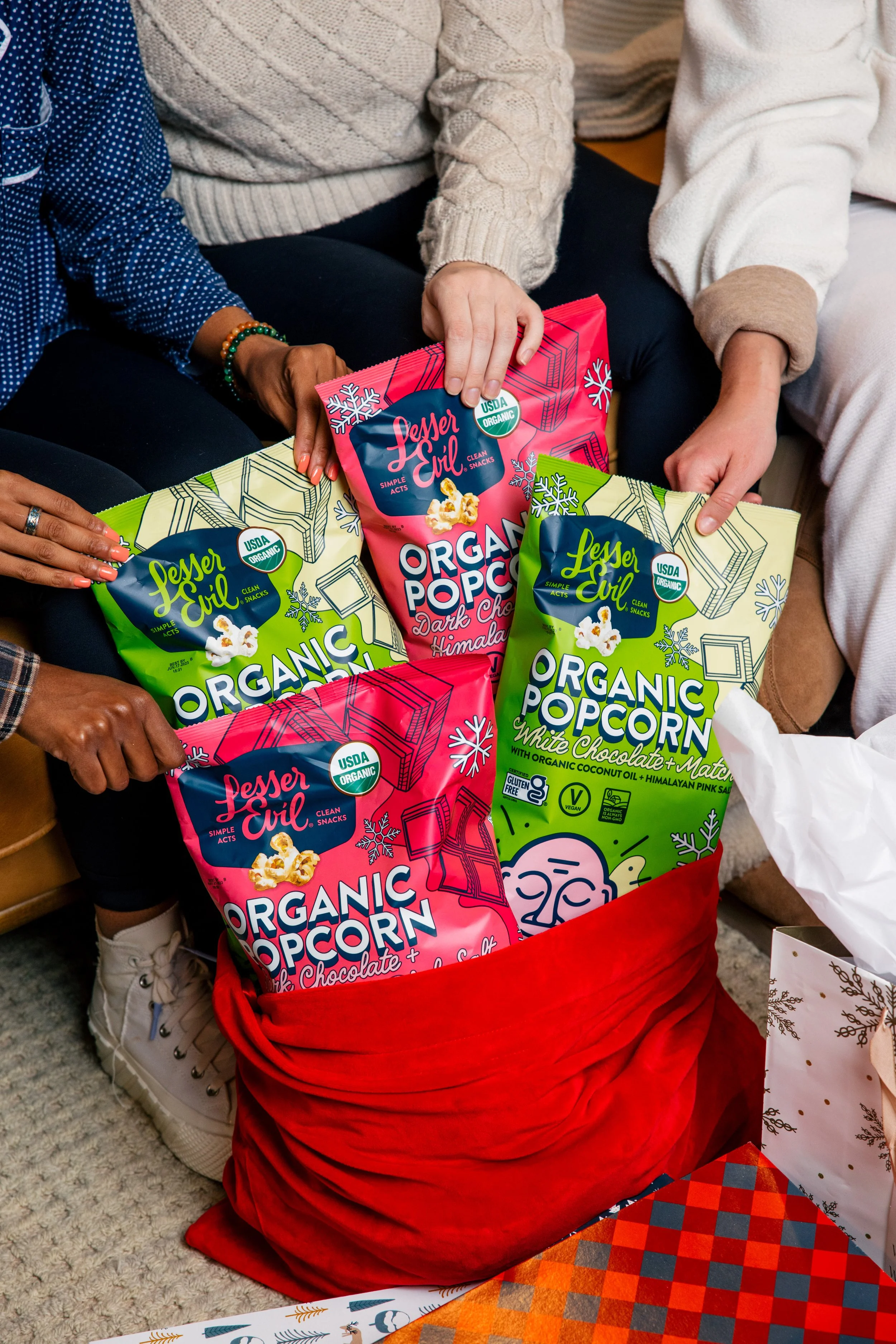 Several individuals gathered around a red bag filled with packages of organic popcorn, including dark chocolate, white chocolate, and regular varieties. The packages feature colorful designs for Commercial product photography Campaign 
