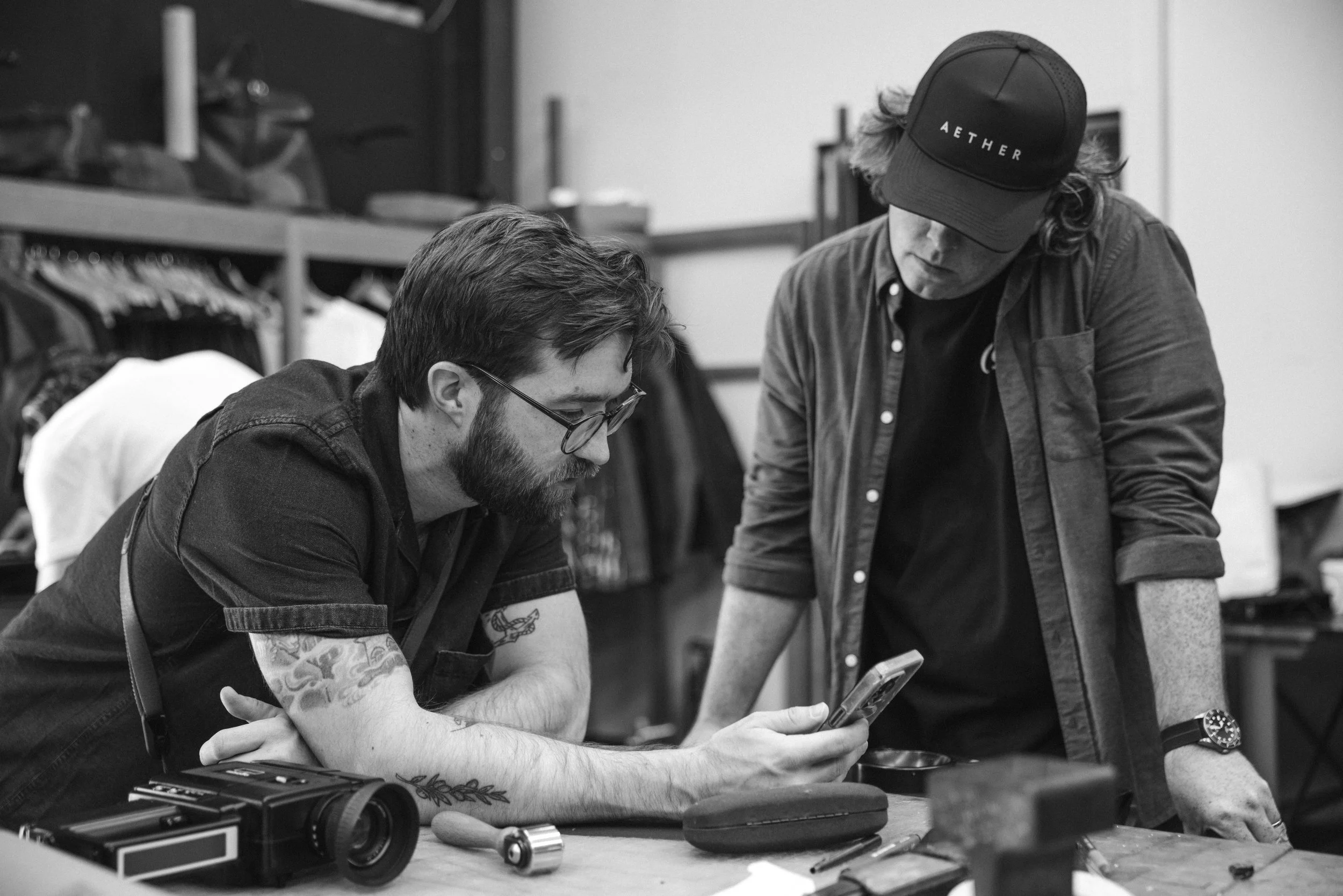 Two men look at a smartphone together on a filmset with various items on a table.